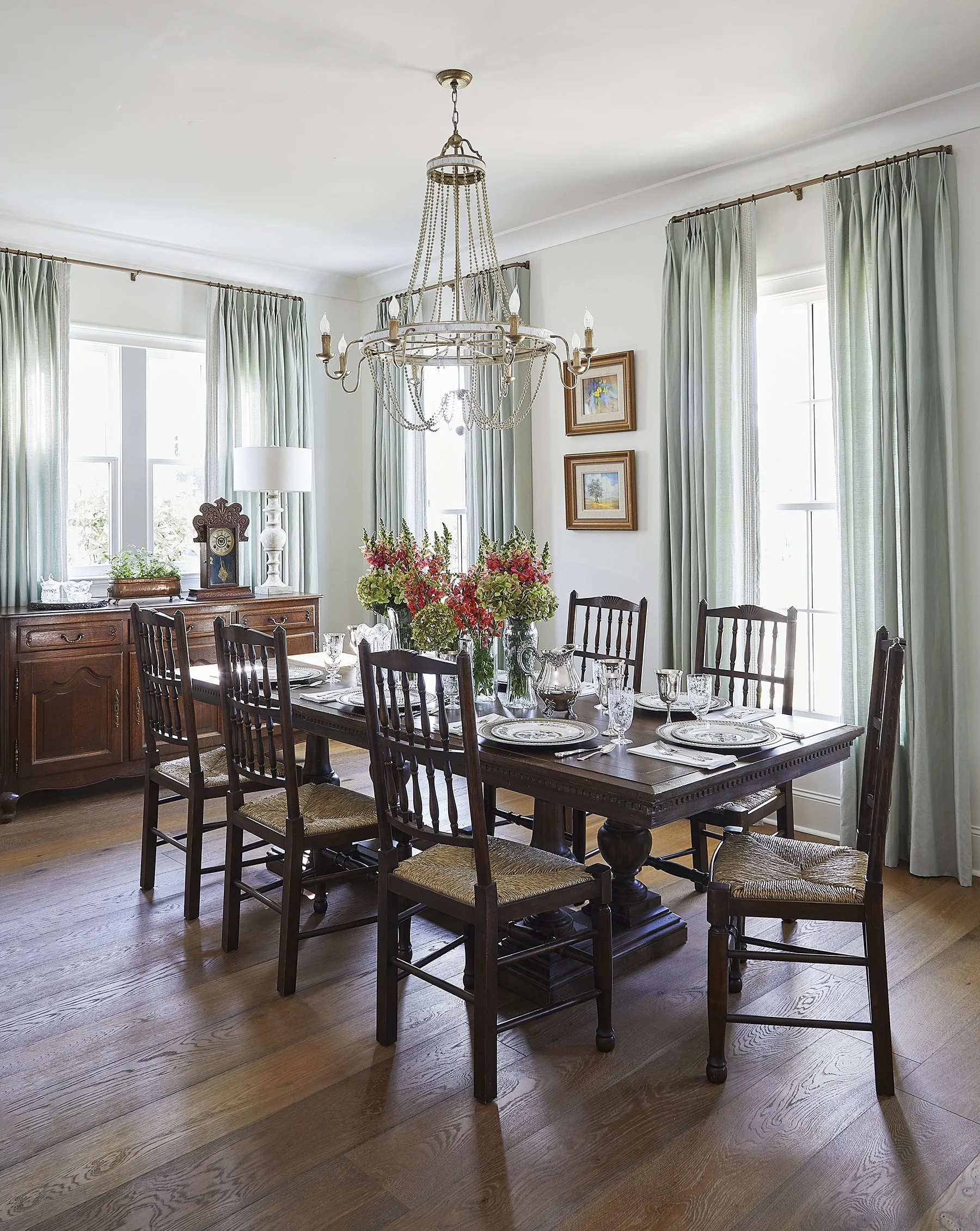 A dining room with a wooden table set with plates, glasses, and a floral centerpiece. There are eight wooden chairs around the table. A chandelier hangs above, and there are windows with light green curtains, a sideboard, a lamp, and framed artwork o