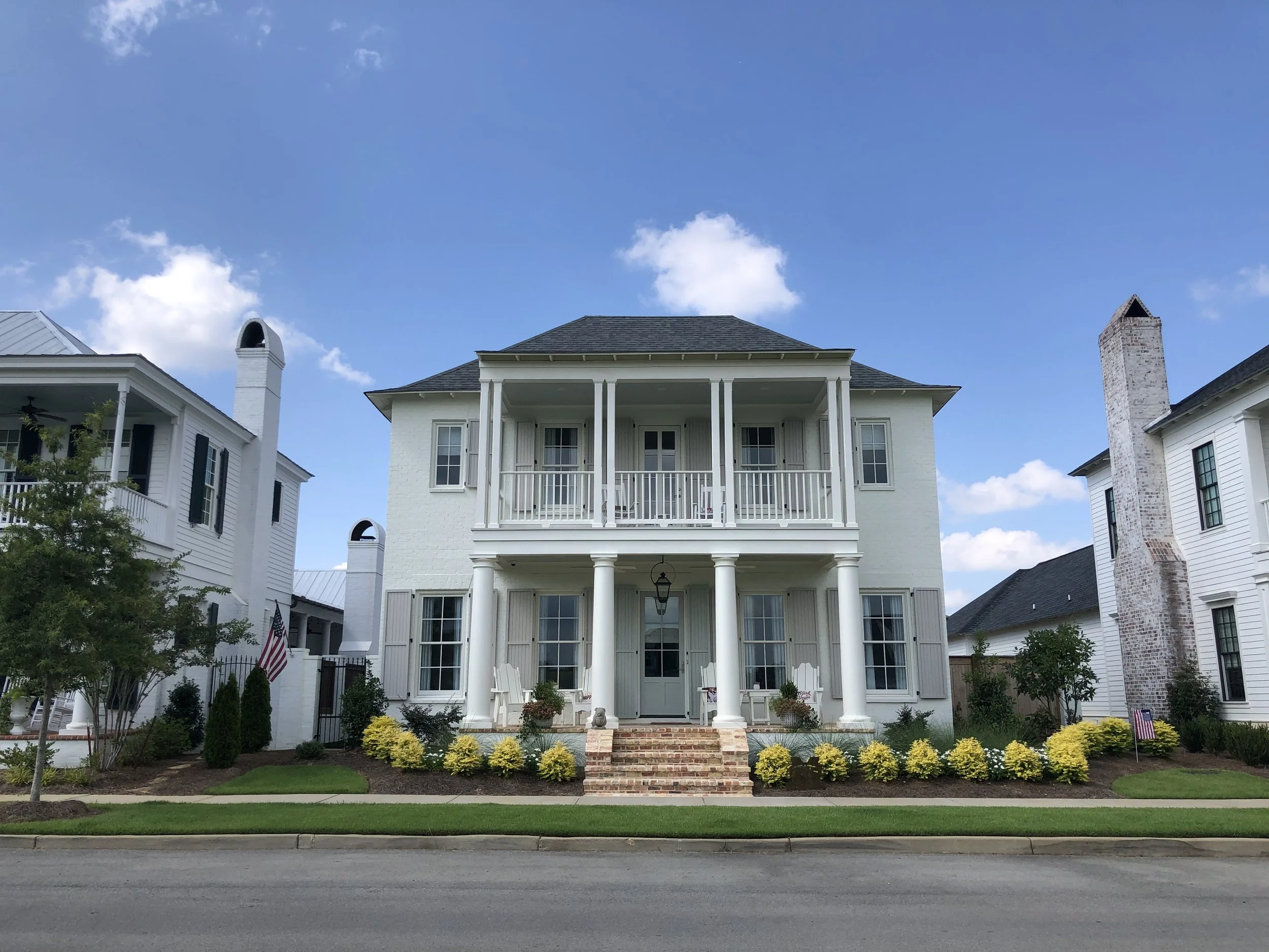 Two-story white house with front porch, columns, and brick steps. Well-maintained lawn with yellow bushes and a small tree.