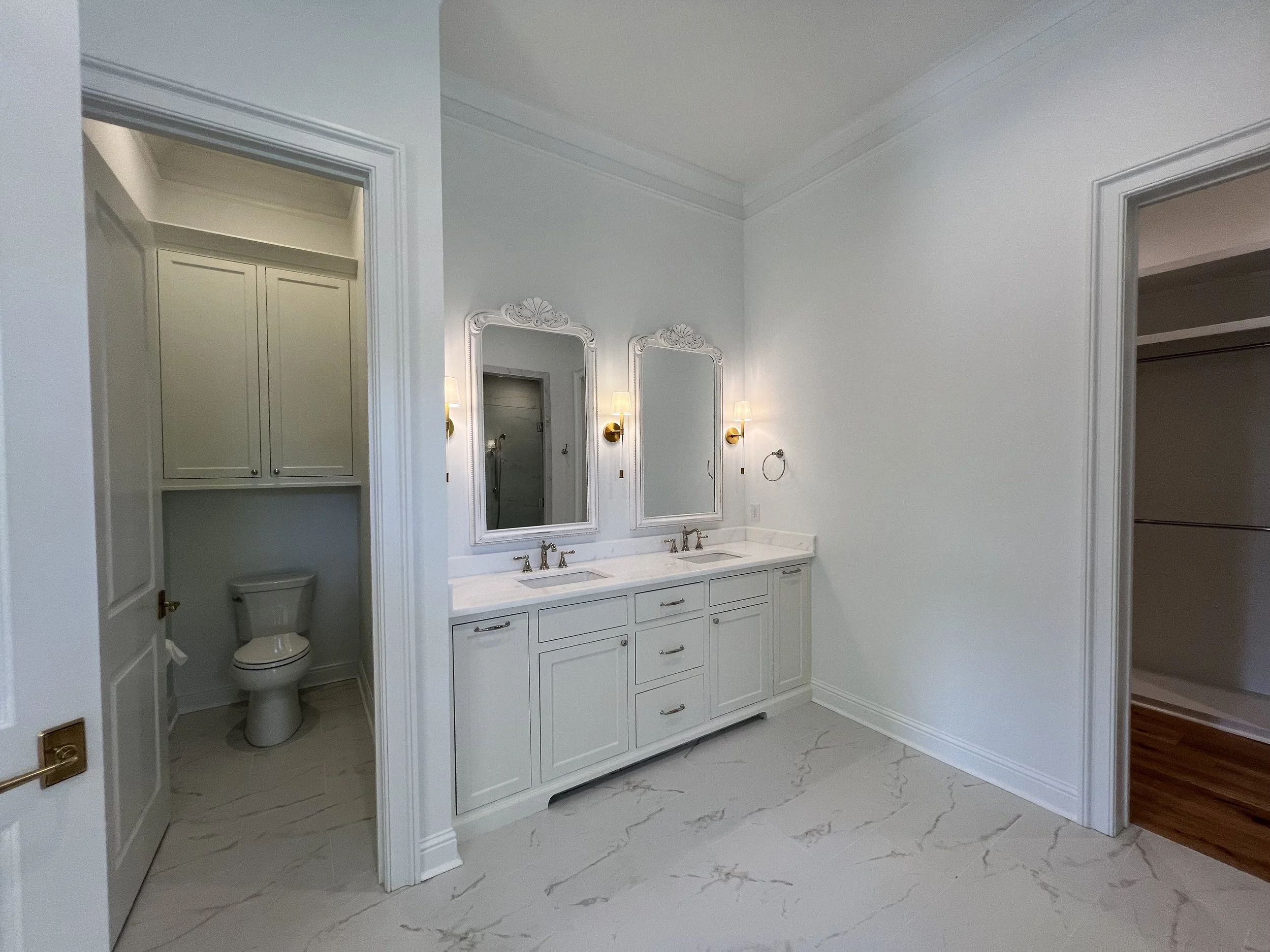 Bathroom with dual vanity sinks, white cabinetry, two mirrors, wall-mounted lights, toilet in a separate small room, and a walk-in closet with wooden flooring.