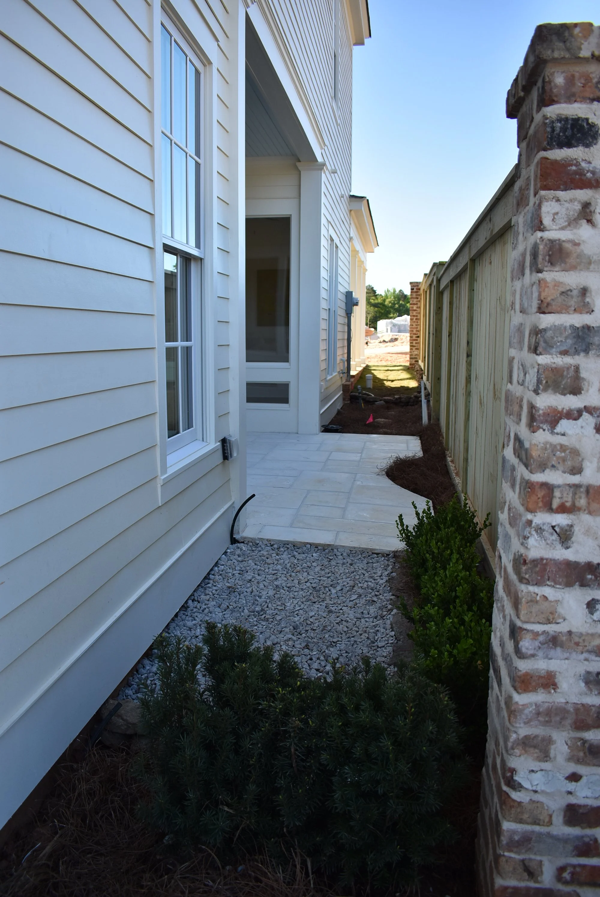 Side view of a house under construction with light yellow siding, a stone pathway, a wooden fence, and some bushes, with clear skies in the background.