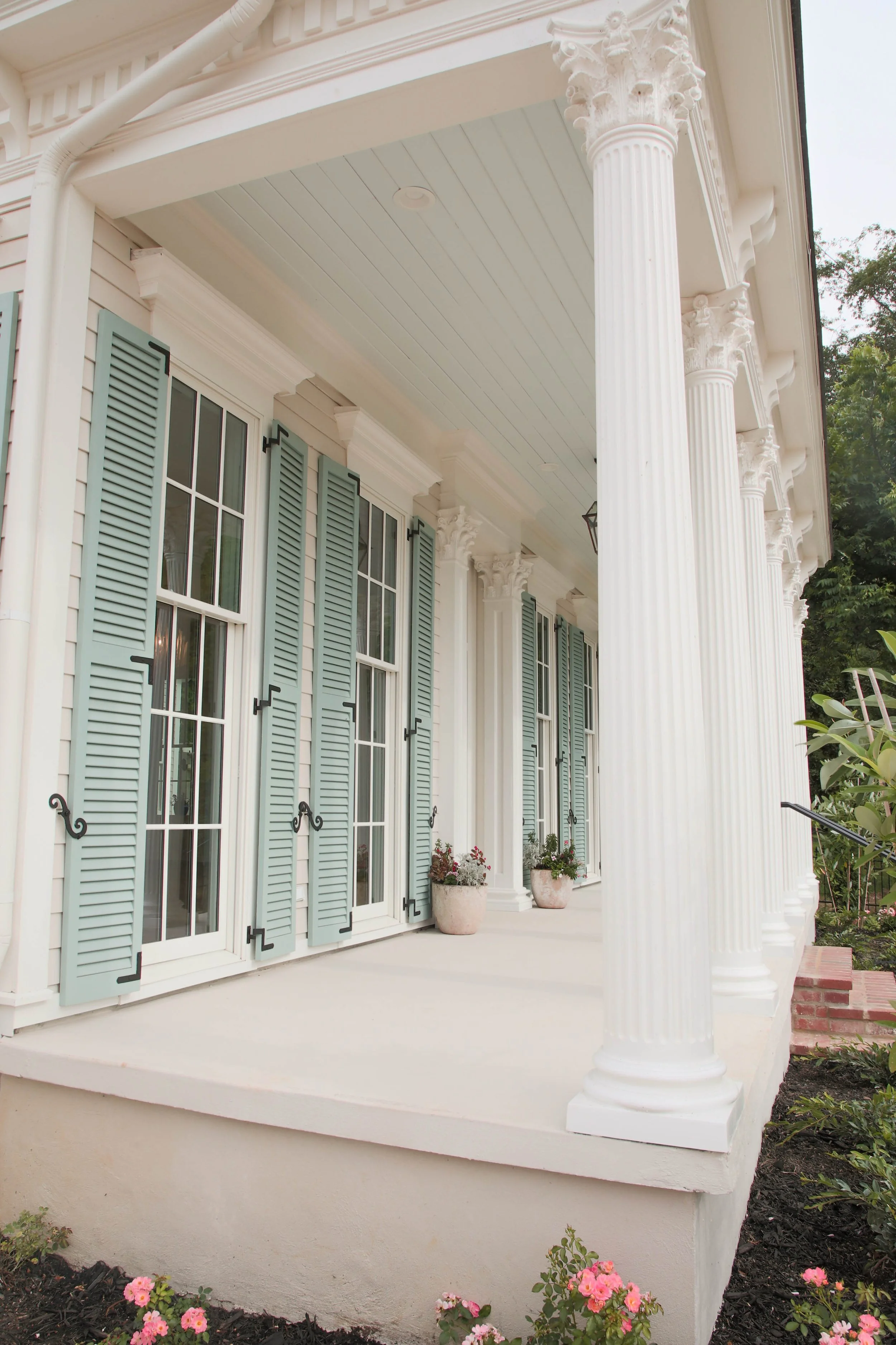 Front porch of a white house with tall columns, light blue shutters, and potted plants, showcasing classical architecture.