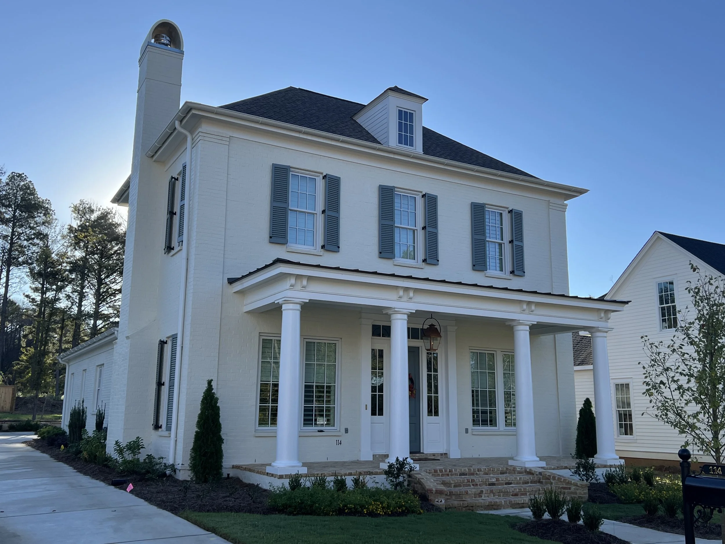 A white, three-story house with black shutters, a covered porch with four white columns, and a brick staircase. There is landscaping with small bushes and trees in front of the house, with a driveway on the left and another white house visible on the