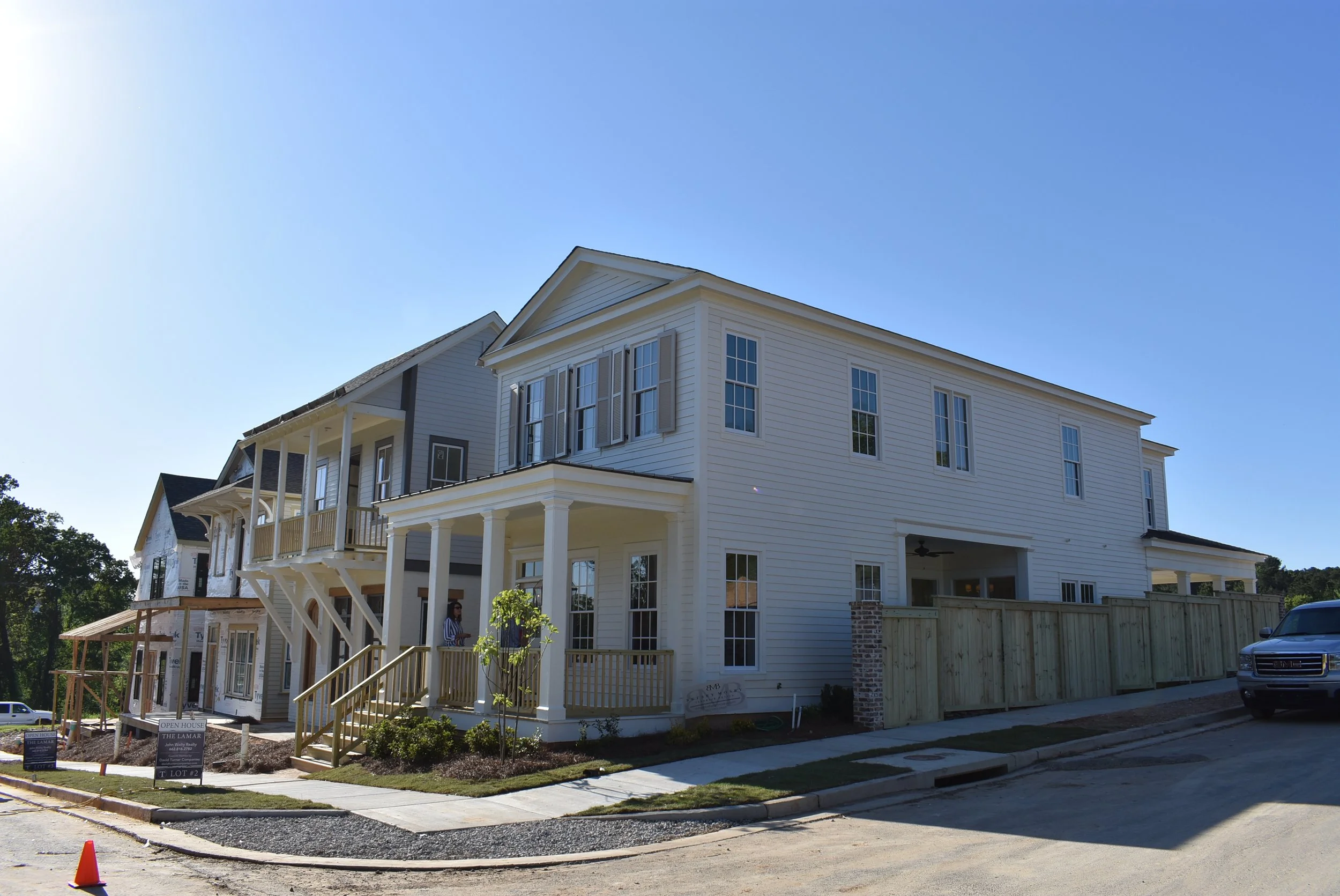 Newly constructed multi-story white house with multiple windows, porches, and balconies, surrounded by a sidewalk, grass, and a construction fence on a sunny day.