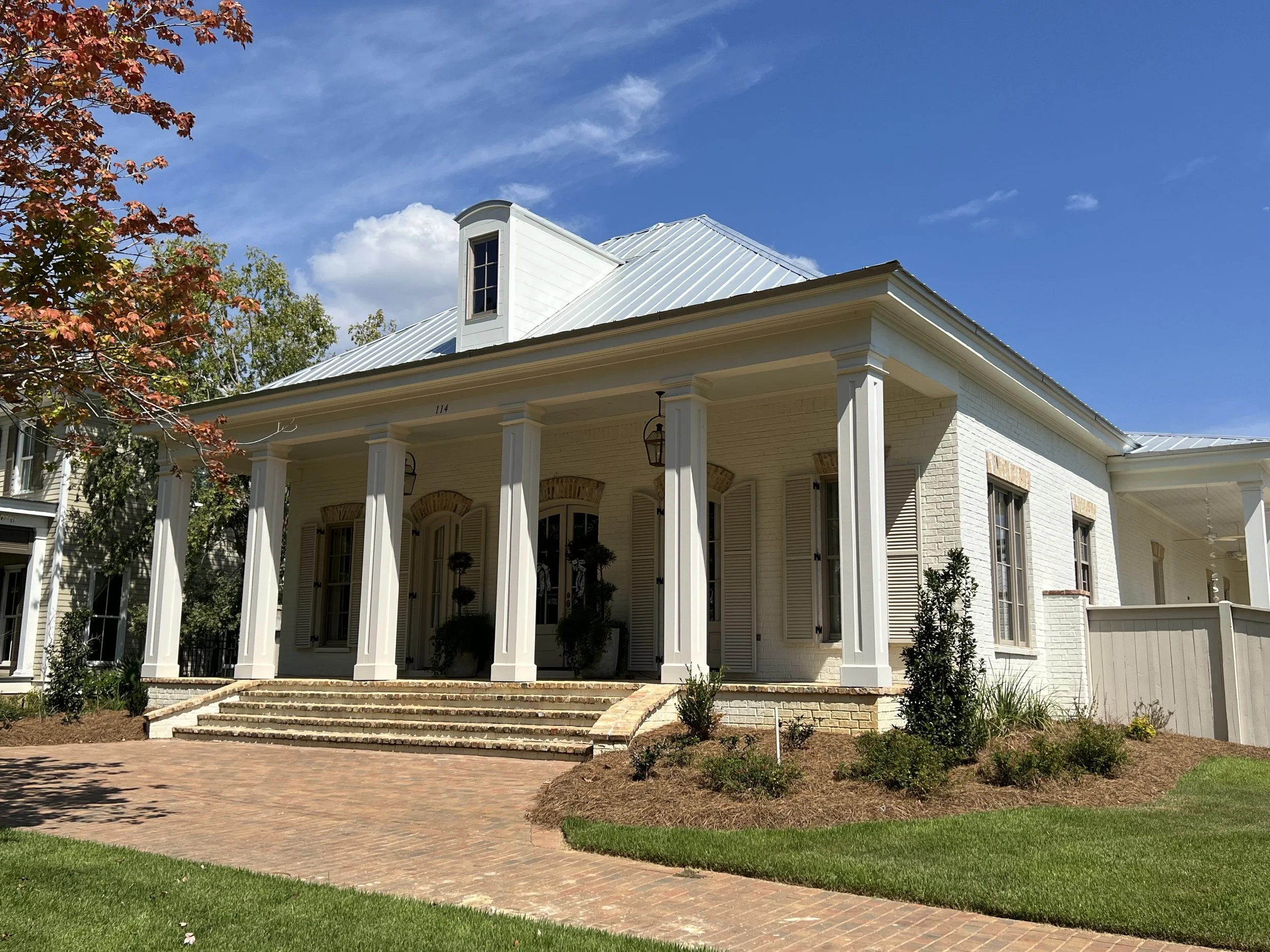 White house with tall columns, brick steps, and a metal roof against a blue sky with some clouds and trees nearby.