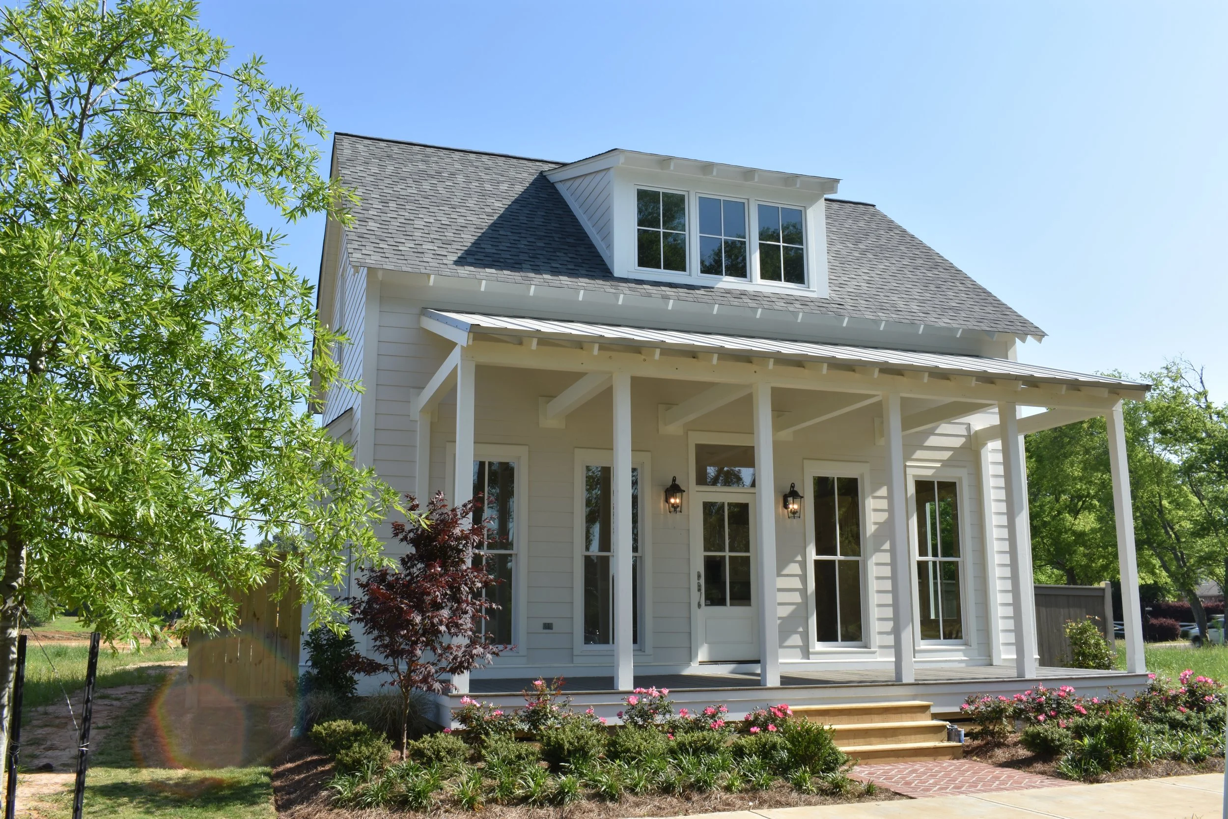 A white two-story house with a gray gabled roof, large front porch, and multiple windows, surrounded by green trees and landscaped garden.