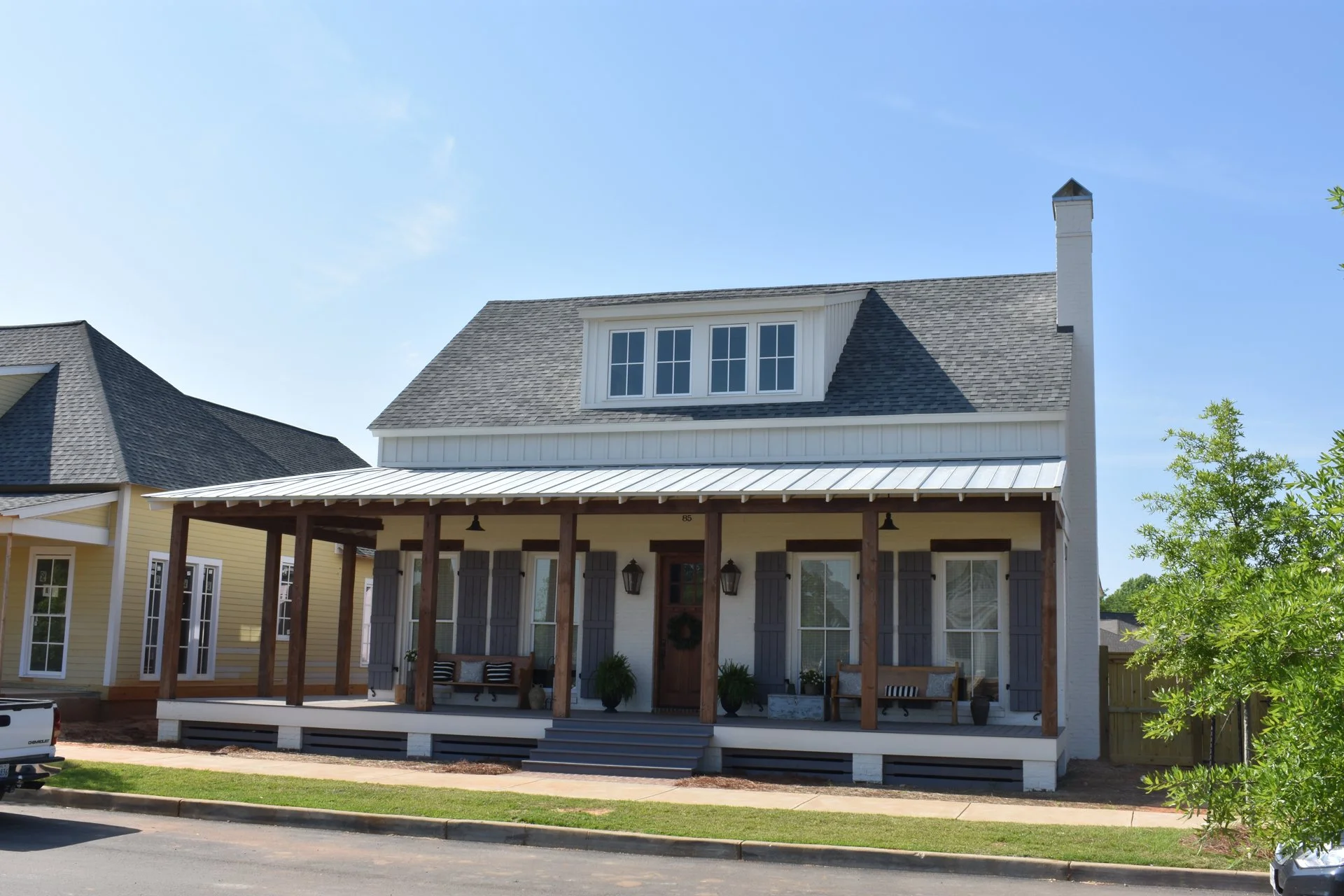 A two-story house with a white exterior, gray shutters, and a metal front porch roof. There are plants and patio furniture on the porch, and a tree is visible on the right side of the house.