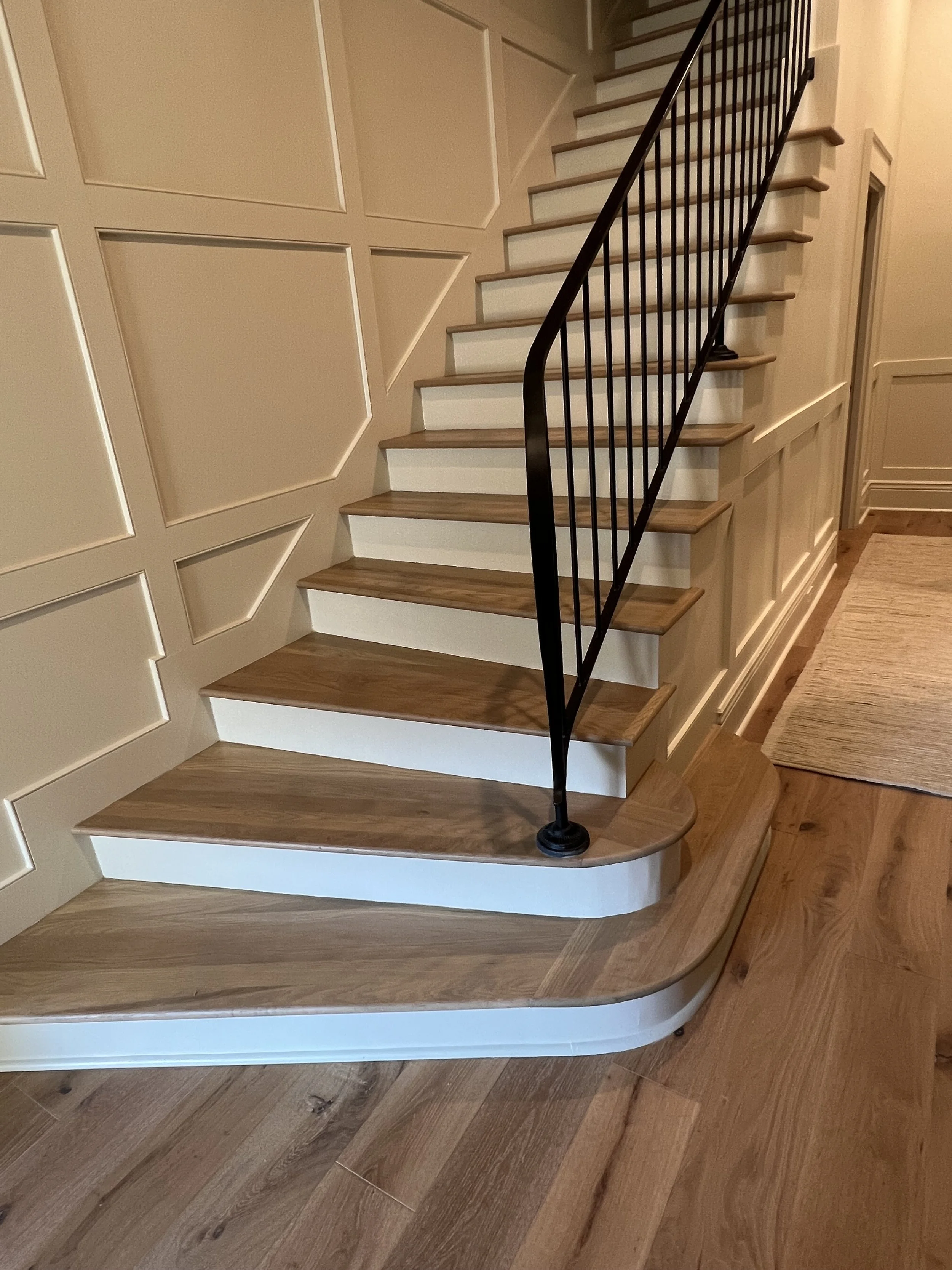 Interior view of a staircase with wooden steps and white risers, beige paneled walls, and a black metal railing, leading up and turning to the right.