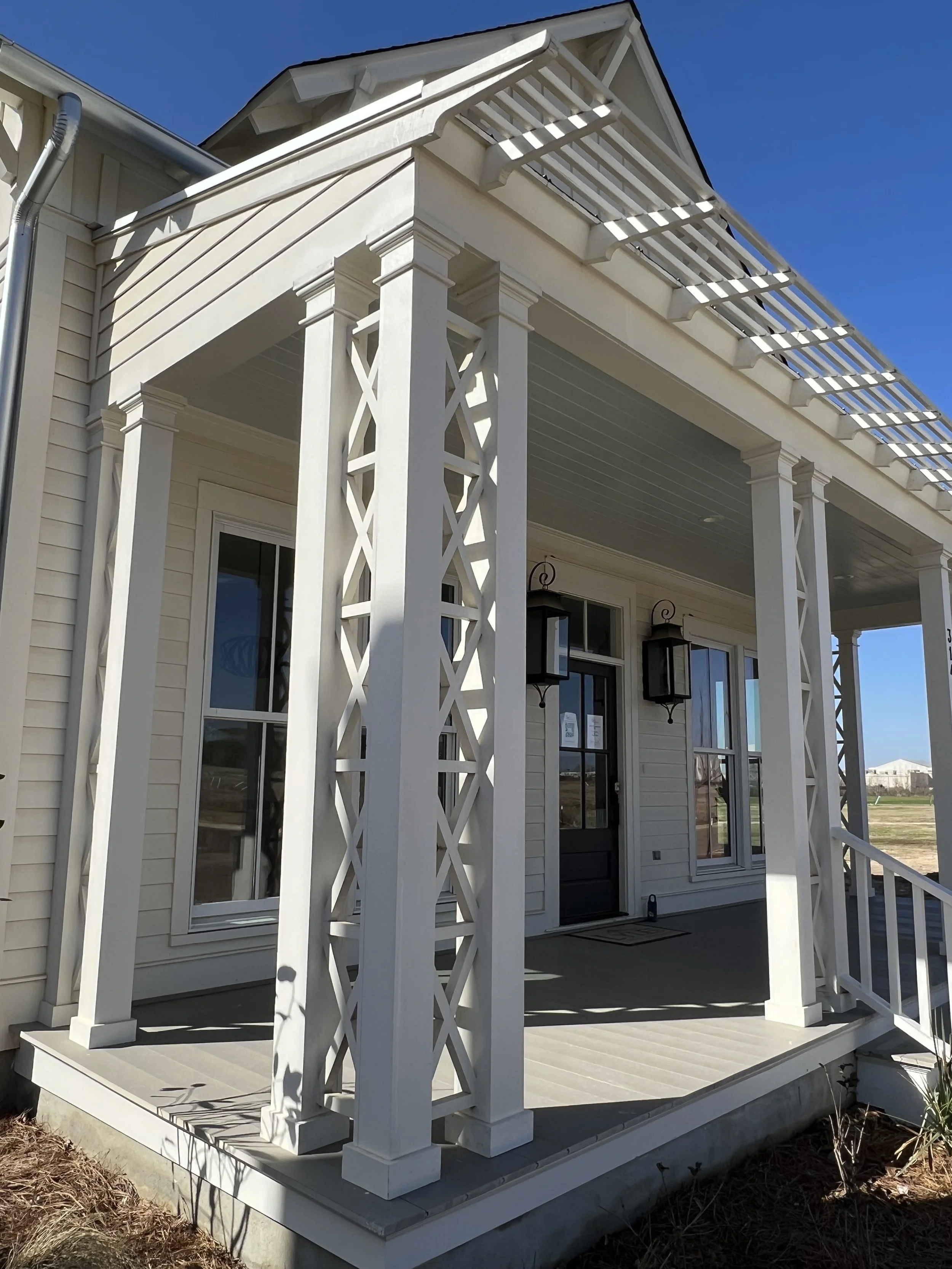 Front porch of a new house with white railings, posts, and lattice detailing, two black lantern-style outdoor lights, and a black front door leading inside.