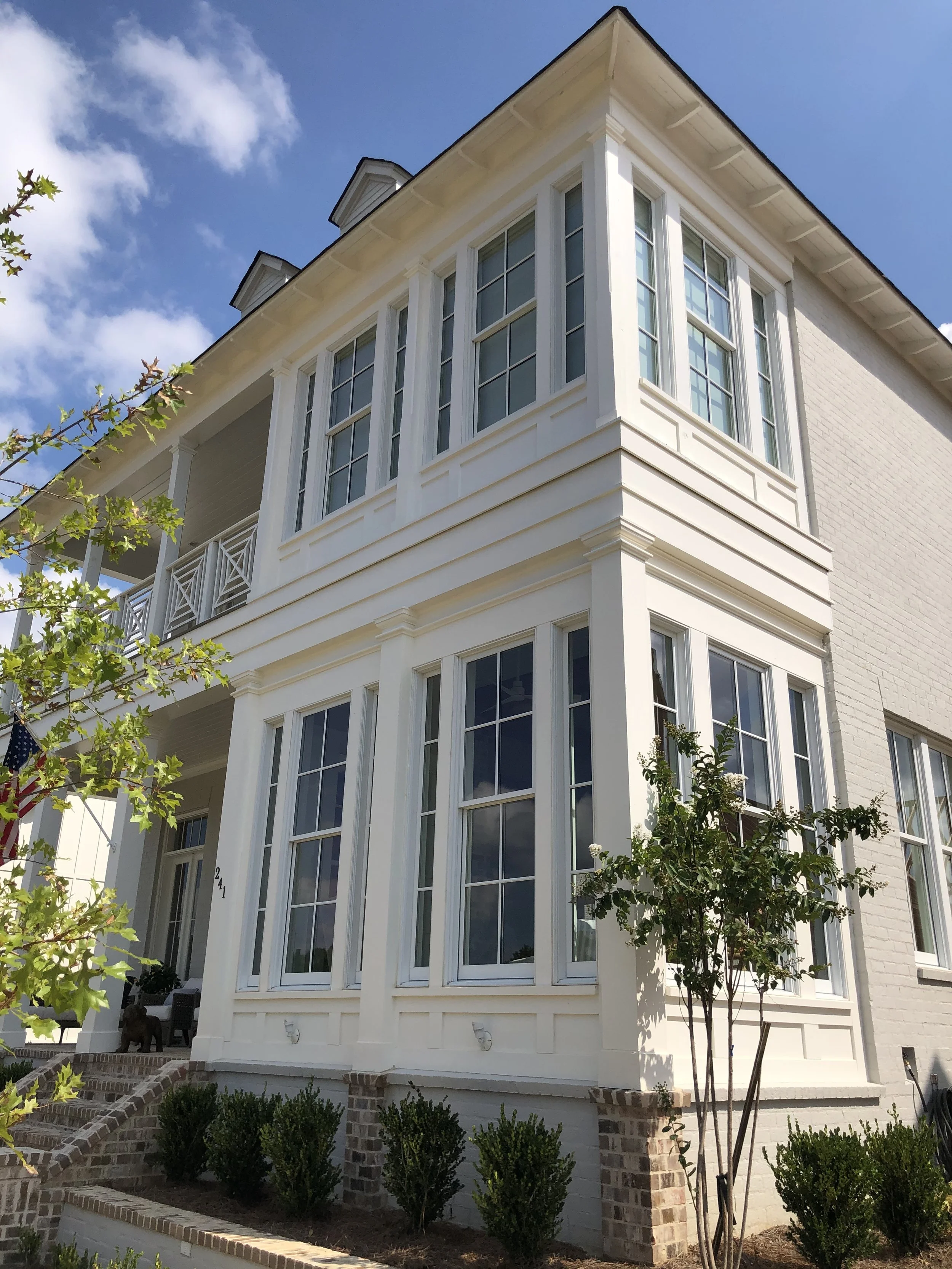 White multi-story house with large windows, brick foundation, and a small front yard with shrubs and young trees, under a blue sky with clouds.