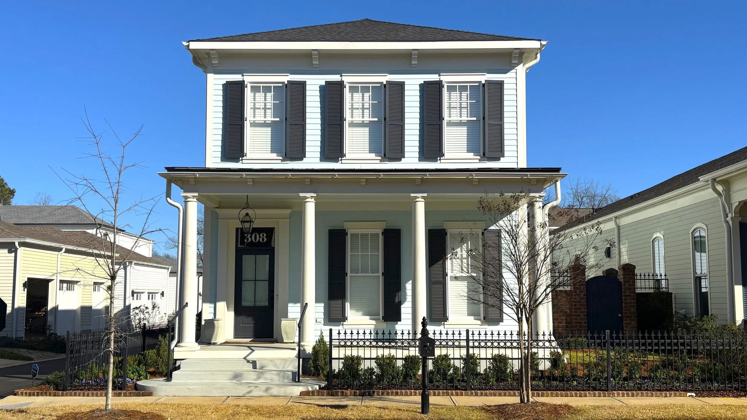 Front view of a two-story light blue house with white trim, black shutters, front porch with white columns, black front door, and a small garden with trees and bushes.
