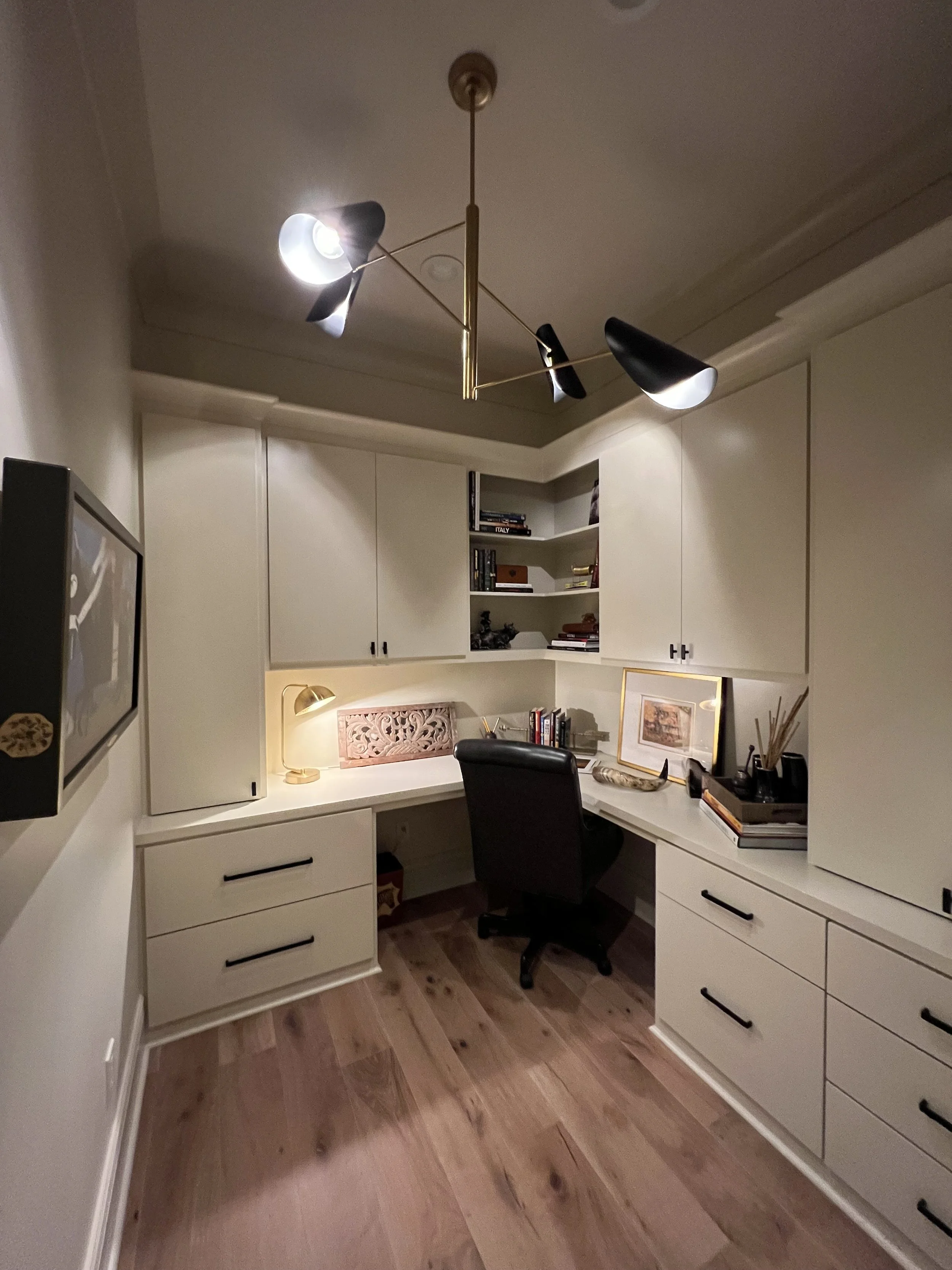 Home office with built-in cream-colored desk and cabinets, black swivel chair, wall-mounted lamp, framed artwork, bookshelf with books, decorative items, modern chandelier, and wooden floor.