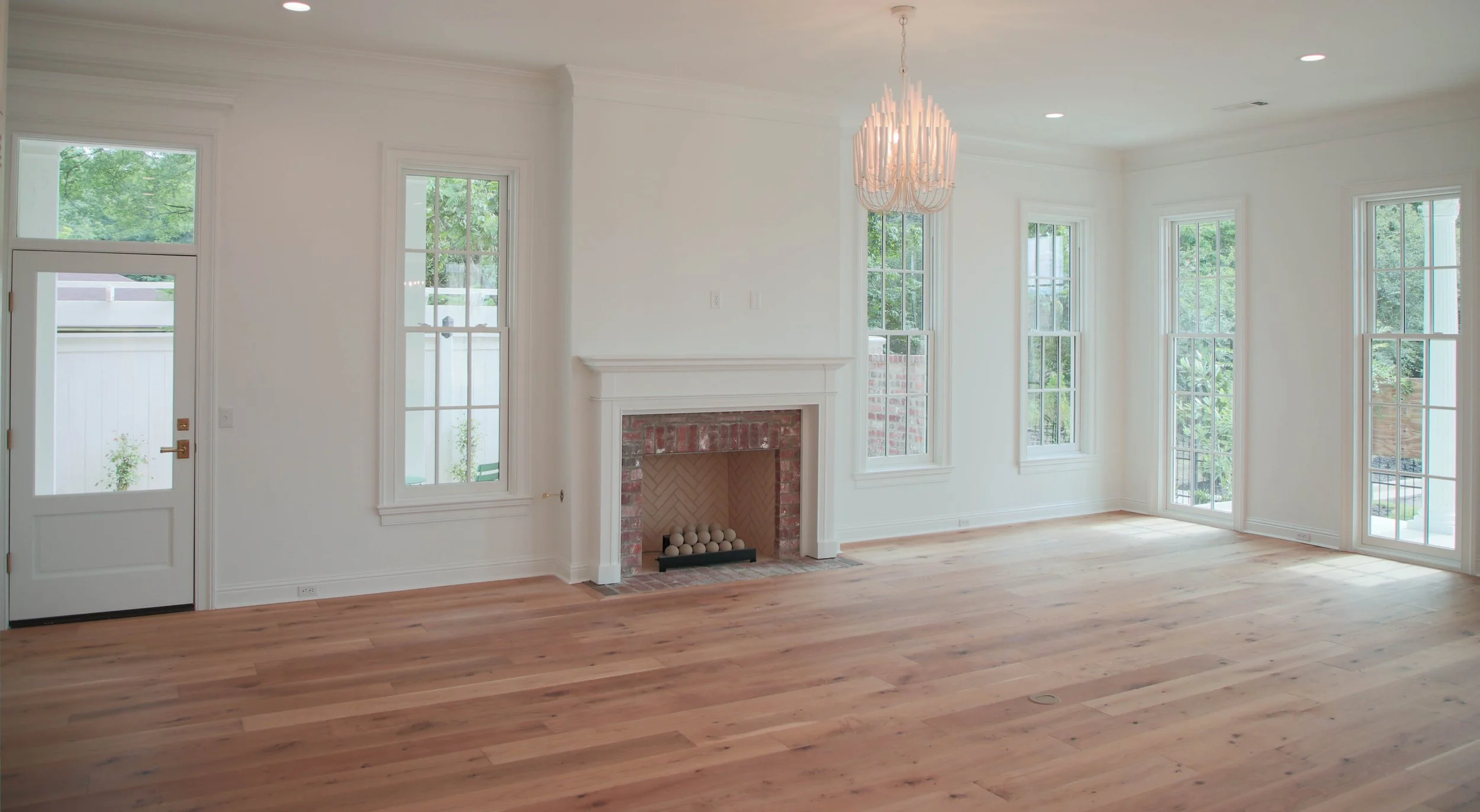 Bright, empty living room with white walls, large windows, a white fireplace with red brick interior, a modern chandelier, and hardwood floors.