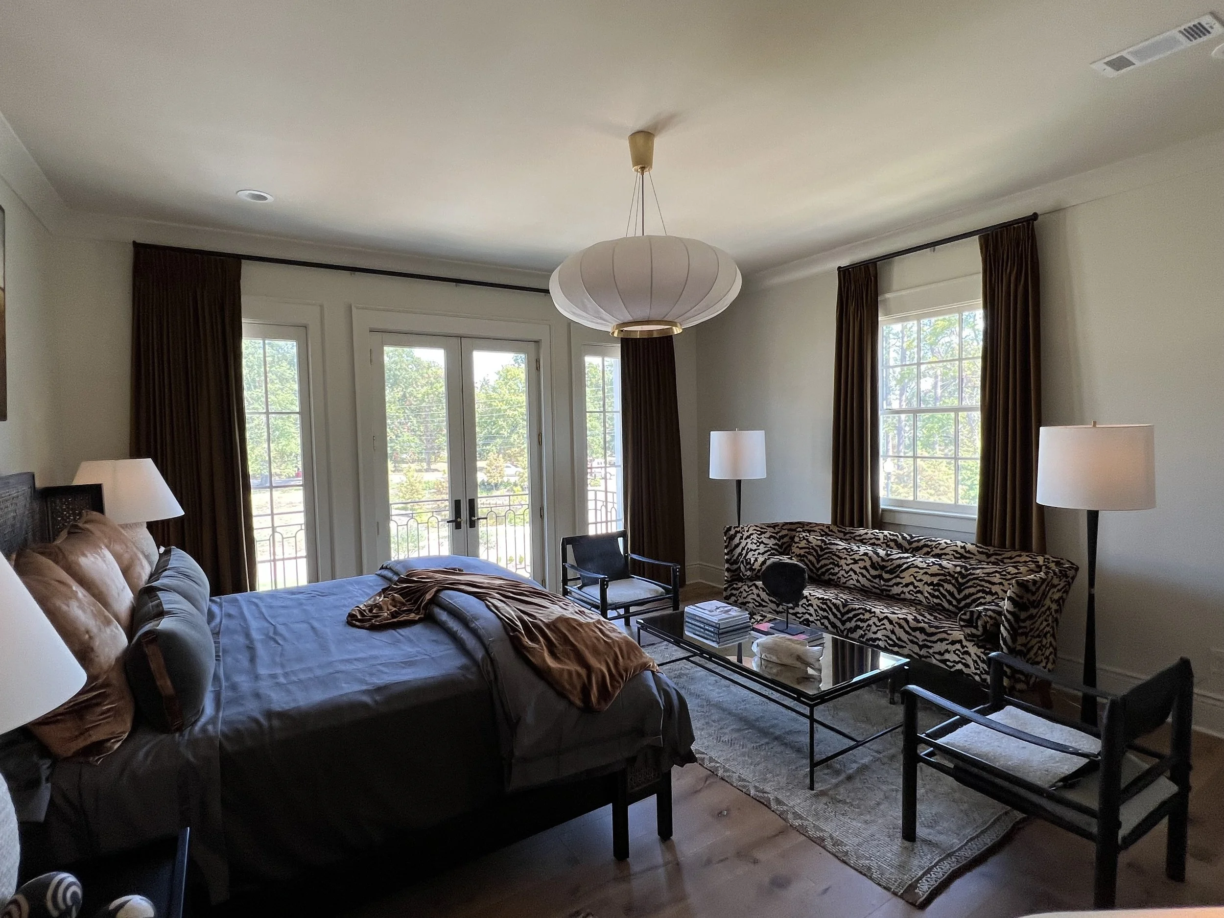 A bedroom with a bed, a sofa with a tiger print pattern, multiple lamps, a glass coffee table, and large windows with brown curtains.