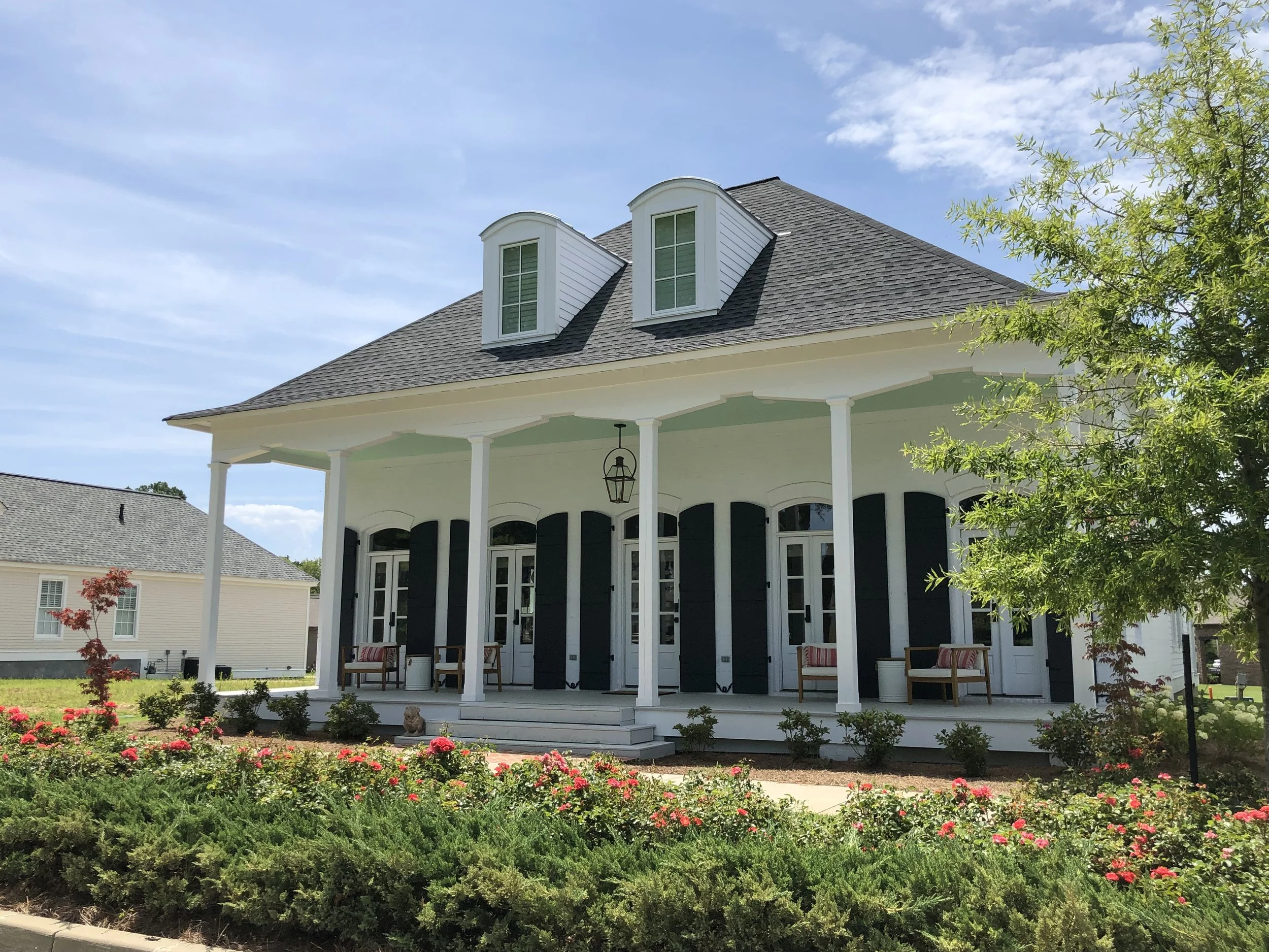 A large white house with a covered front porch, black shutters, and dormer windows on the roof. The porch has two benches, a hanging lantern, and is surrounded by greenery and flowers.