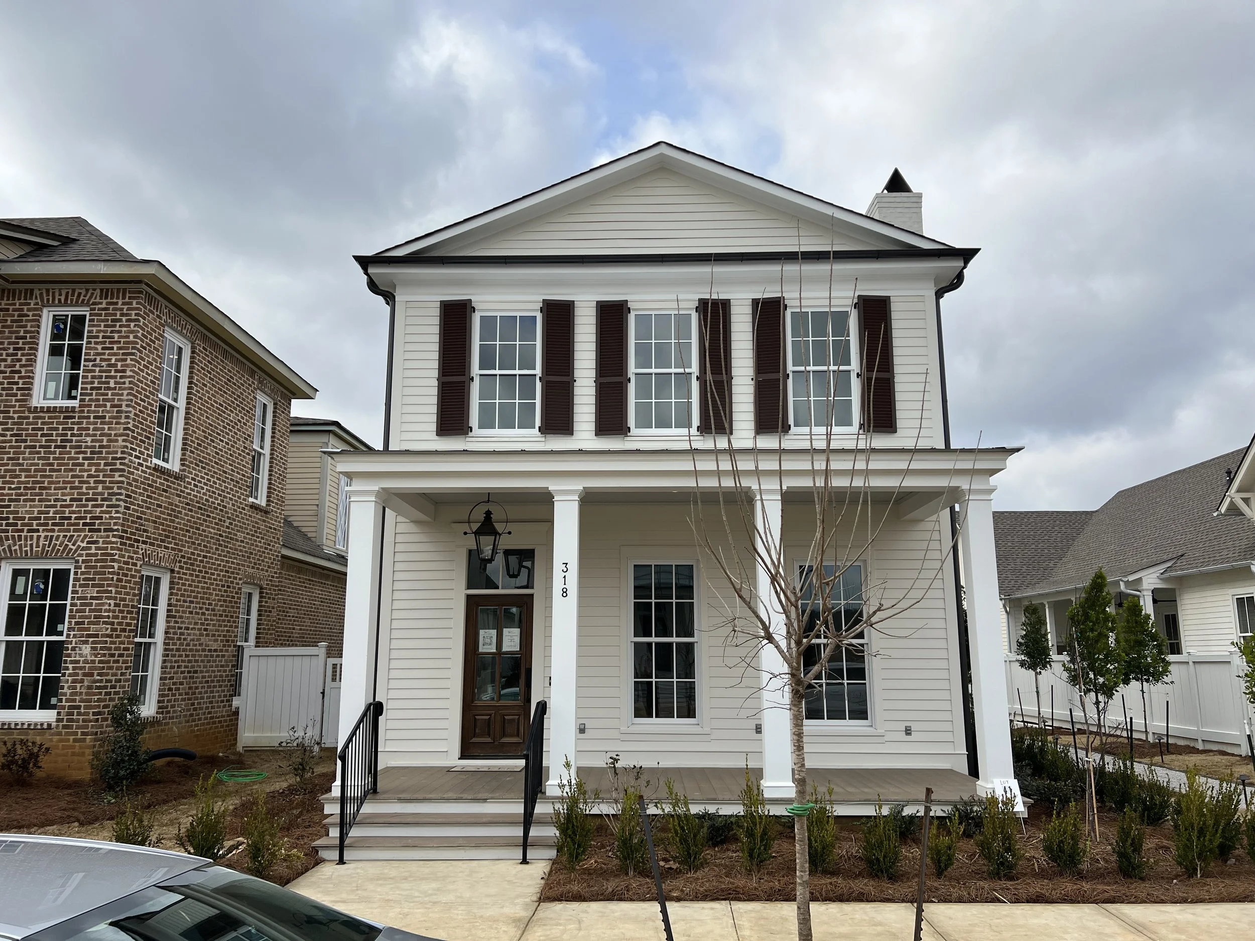 Front view of a two-story white house with brown shutters, a porch, and a leafless tree in front, neighboring houses on either side, under a cloudy sky.