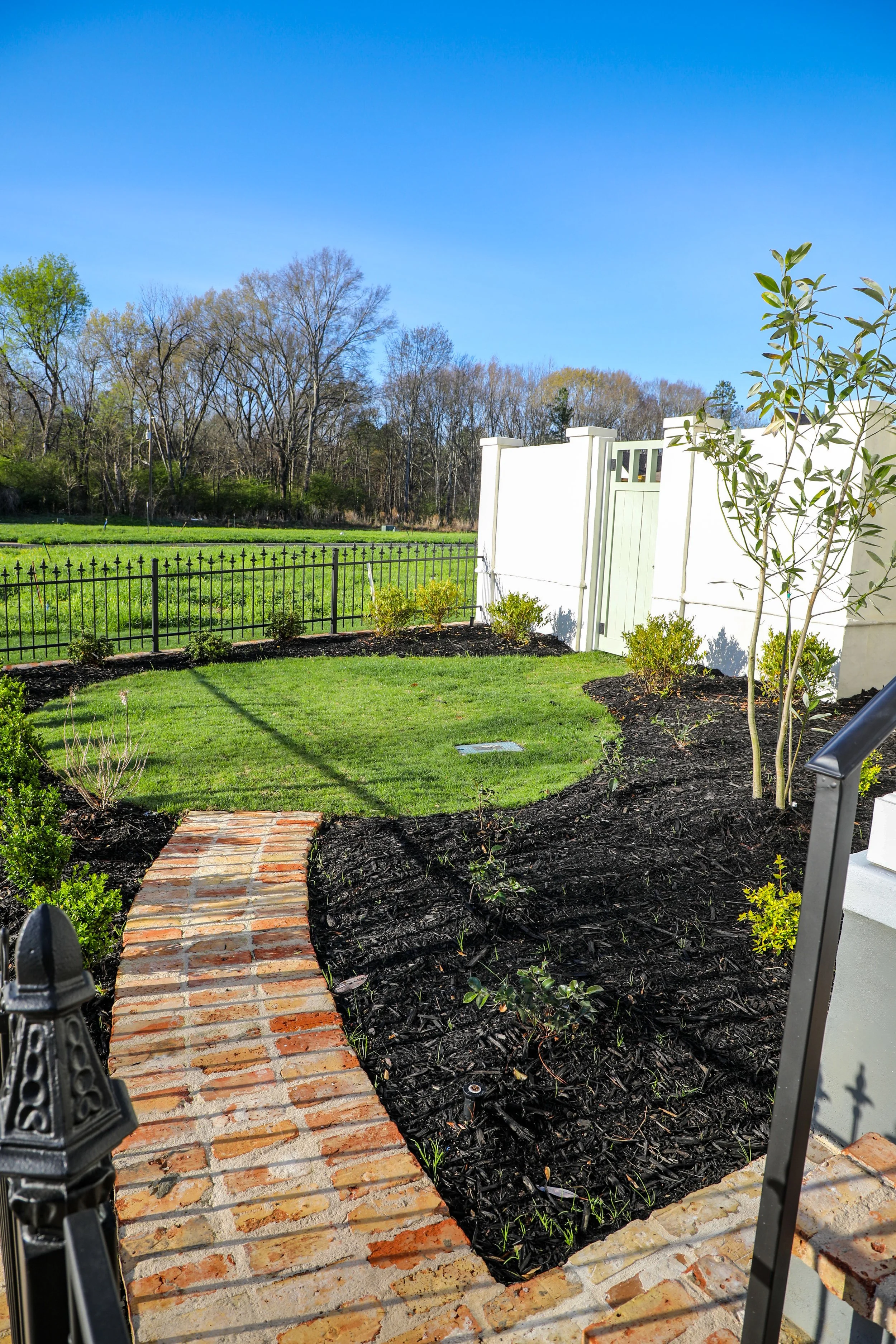 A landscaped backyard with a brick pathway, green grass, small shrubs, a white fence gate, and trees in the background under a clear blue sky.