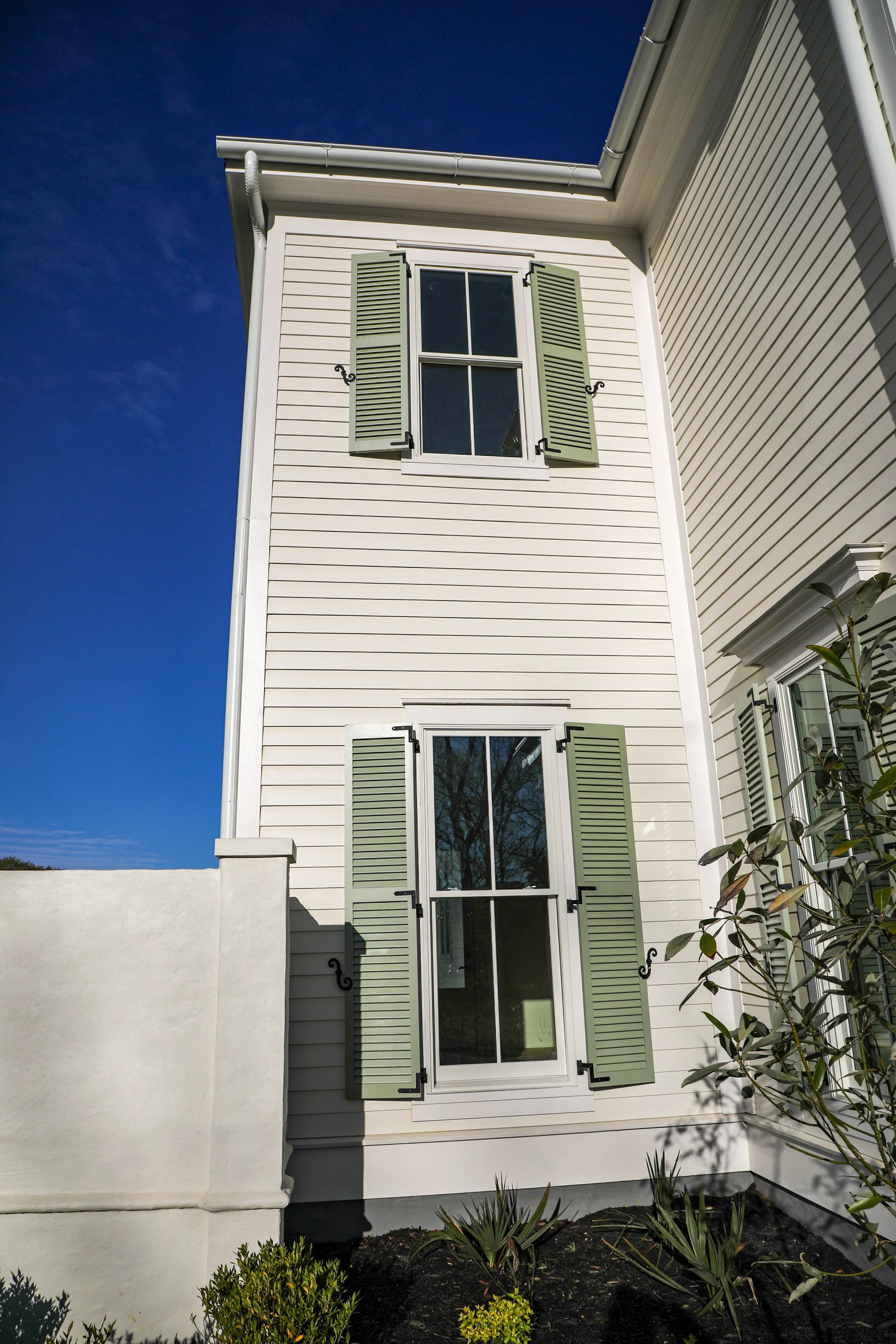 Exterior view of a white house with two large windows with green shutters, taken on a clear sunny day with a blue sky.