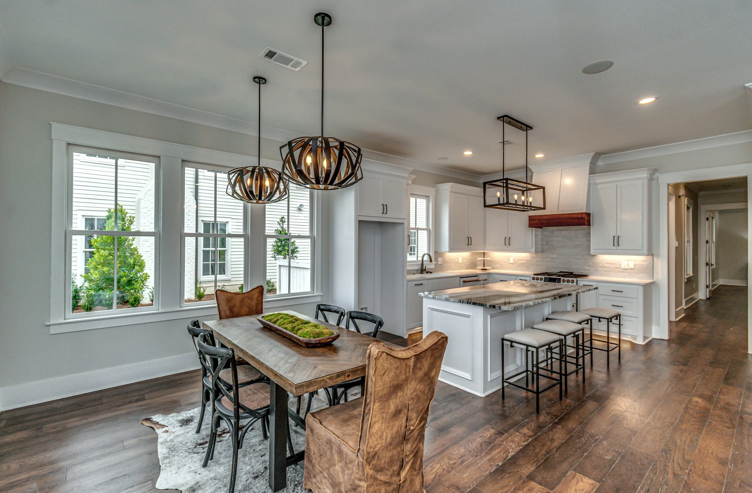 Open kitchen and dining area with white cabinetry, dark wood floors, and unique pendant lighting fixtures. The dining table has a mix of chairs, including two brown leather armchairs and black metal chairs. Large windows provide natural light and a v