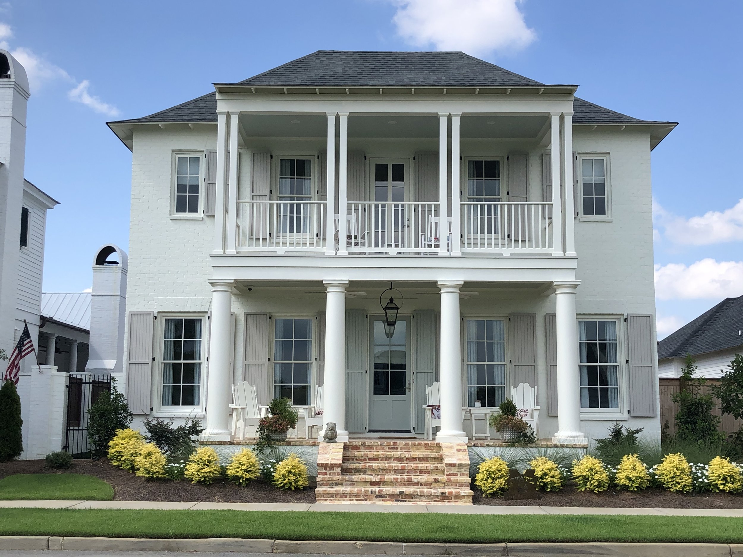 White two-story house with shutters, front porch with columns, and landscaping with shrubs and yellow flowers.