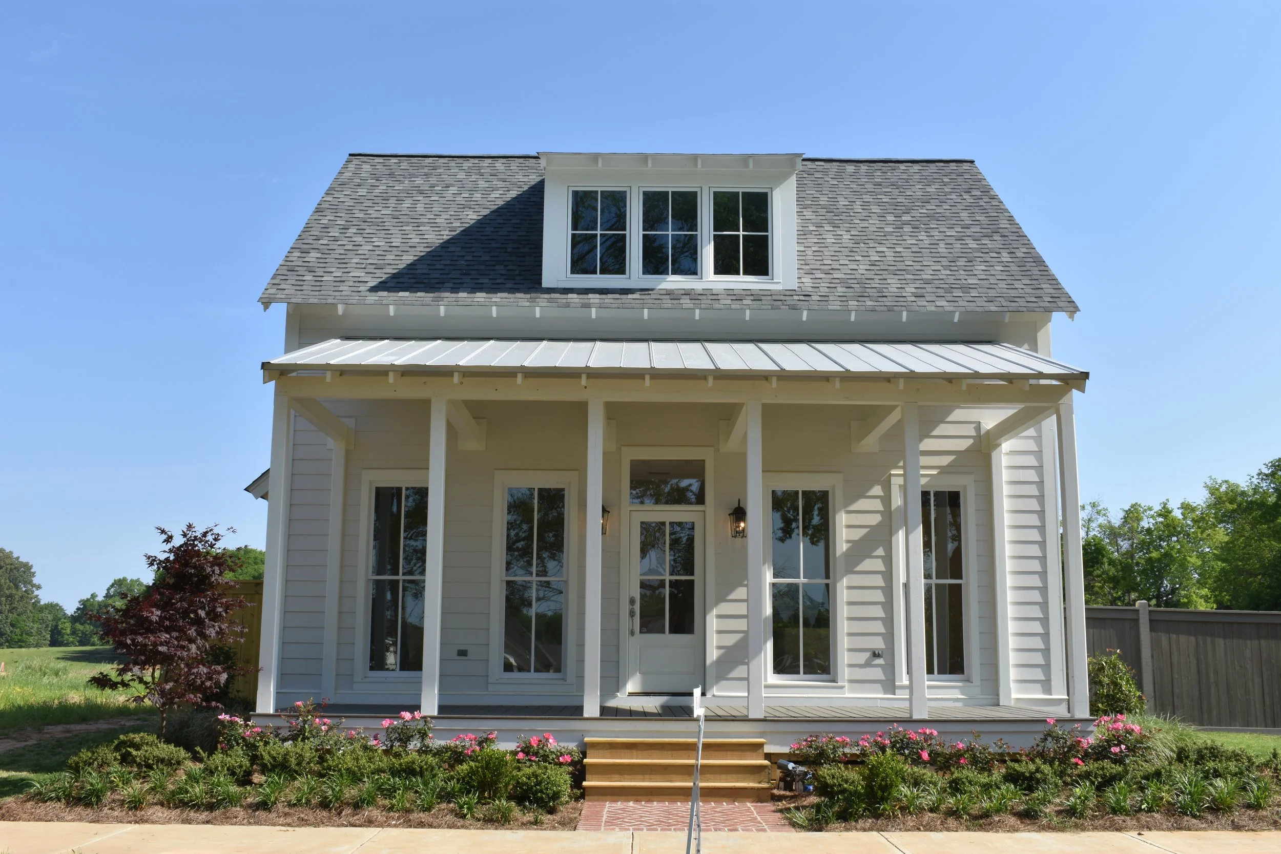 Front view of a modern two-story house with white siding, a gabled roof, large windows, and a porch with white columns, surrounded by a garden with pink flowers and green shrubs.