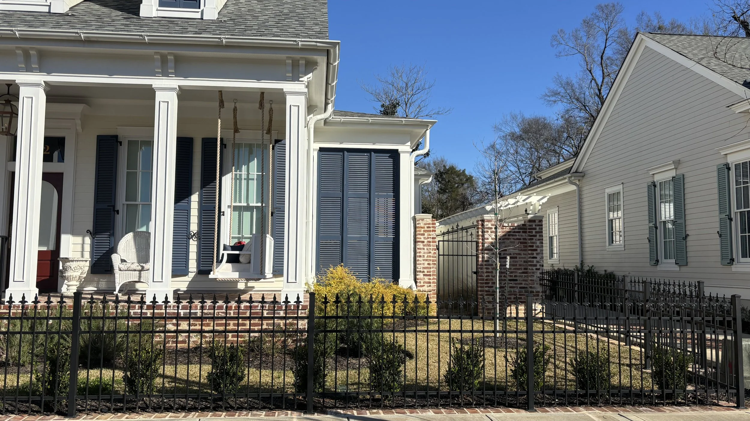 Front view of two neighboring houses with a small outdoor porch, white and cream siding, window shutters, a black metal fence, and a vegetable garden with small bushes and a young tree.