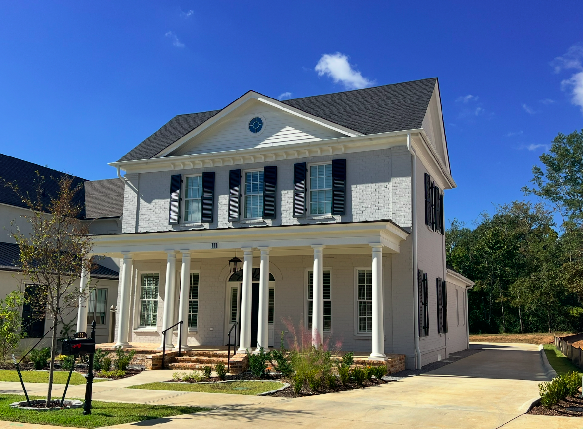 White two-story house with black shutters, front porch with white columns, black mailbox, and a driveway, under a blue sky with a few clouds.