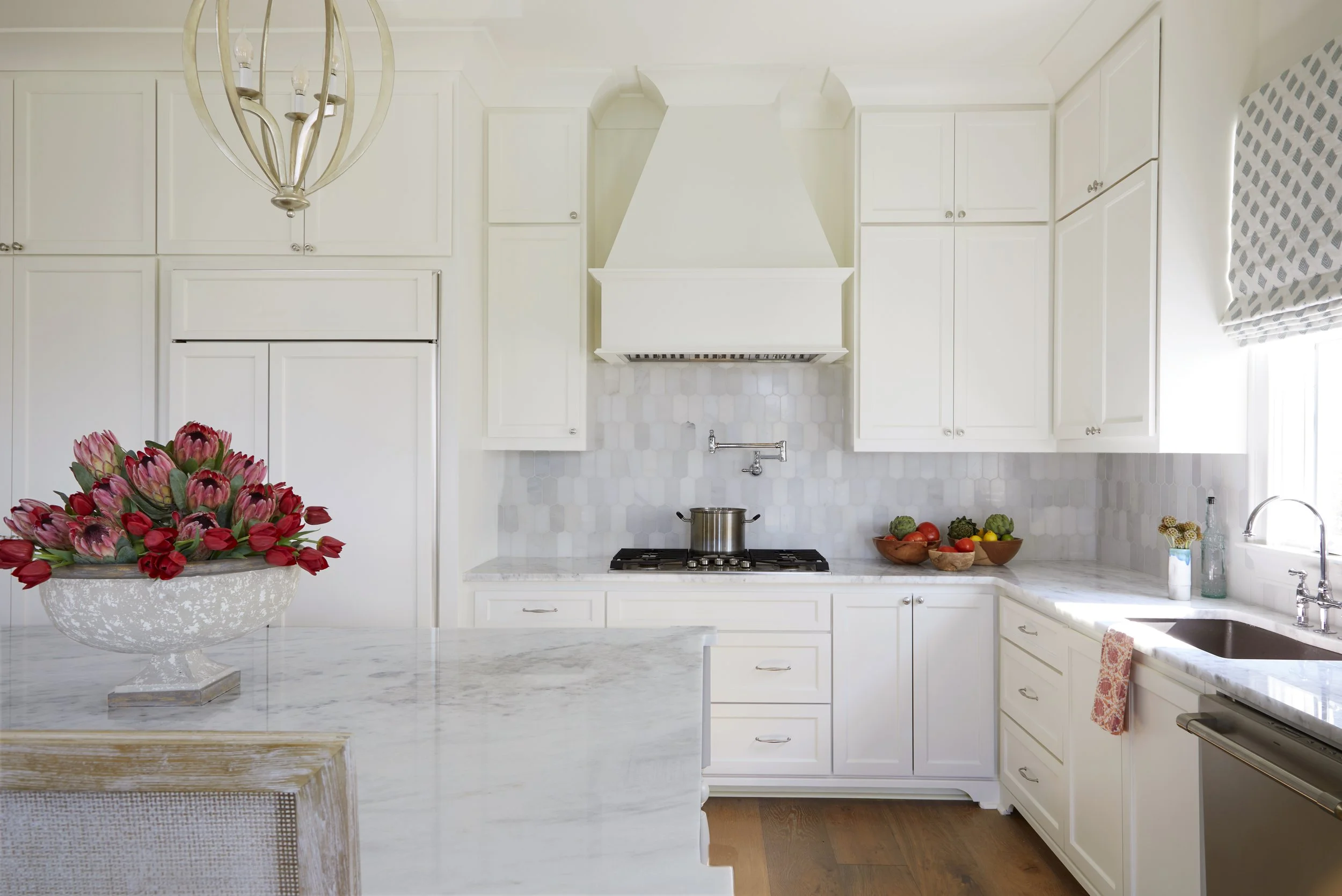 Bright white kitchen with marble countertops, white cabinets, a chimney range hood, and a window with a patterned shade. There are bowls of vegetables on the counter and a flower arrangement in a decorative vase.