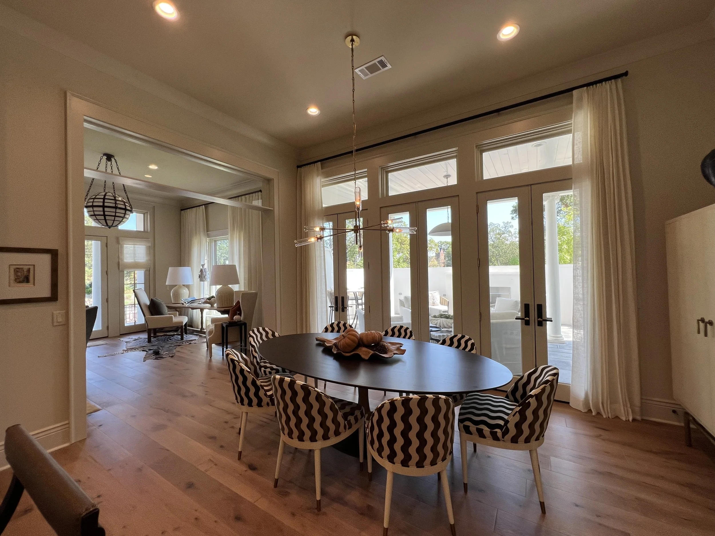 Interior view of a dining area with a round table and striped chairs, leading to a sunlit outdoor patio through large glass doors.