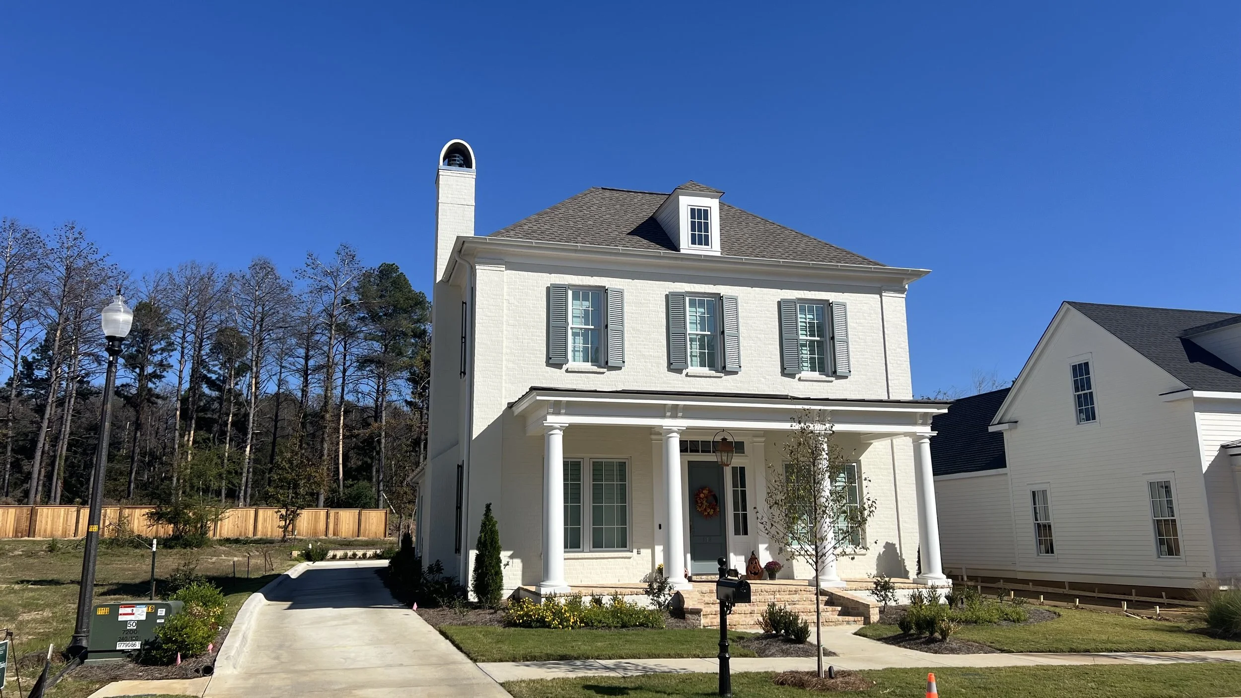 White two-story house with a porch, columns, and shutters, surrounded by a well-kept lawn and trees, under a clear blue sky.