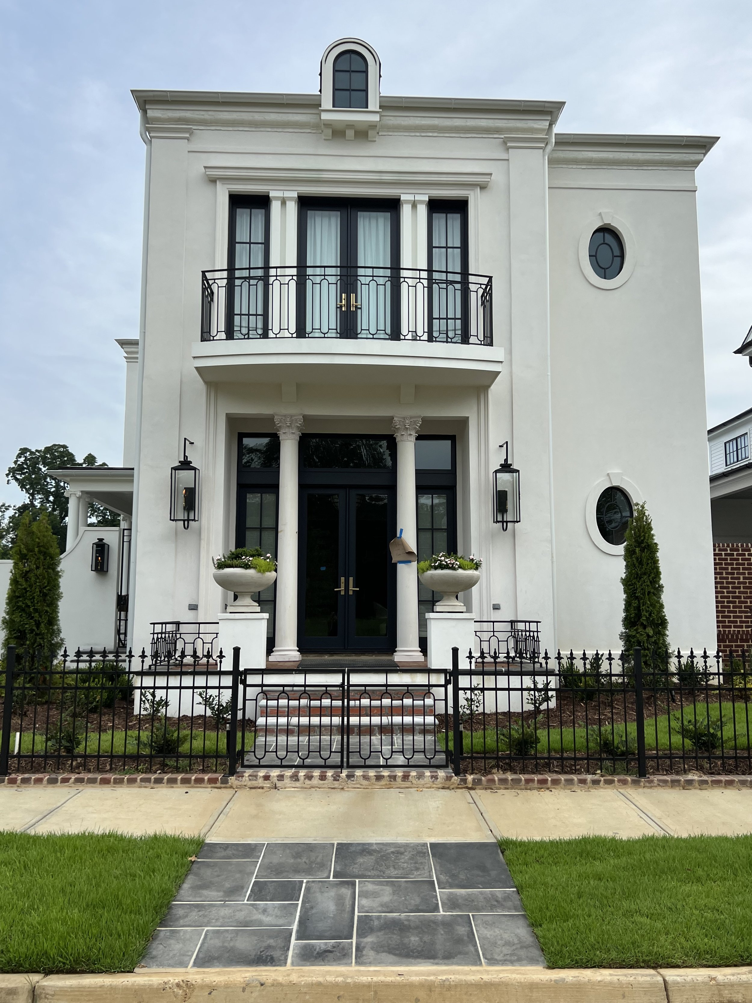 Front of a white multi-story house with a small balcony, black doors, and decorative columns at the entrance, surrounded by a small black metal fence and green lawn.