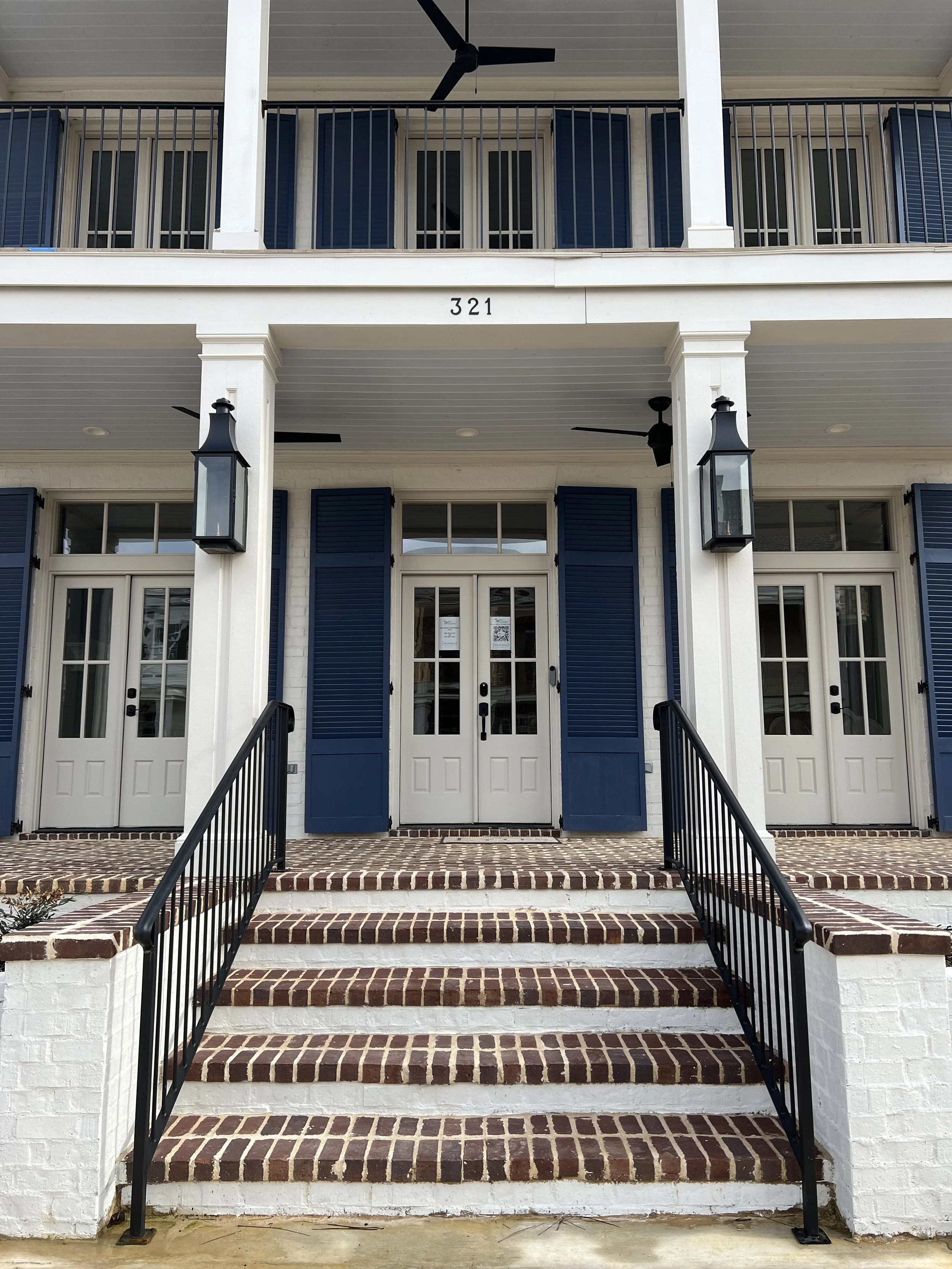 Exterior view of a white multi-story residential building with blue shutters, black lantern-style lights, a staircase with black handrails, and a balcony with ceiling fans. The building number is 321.