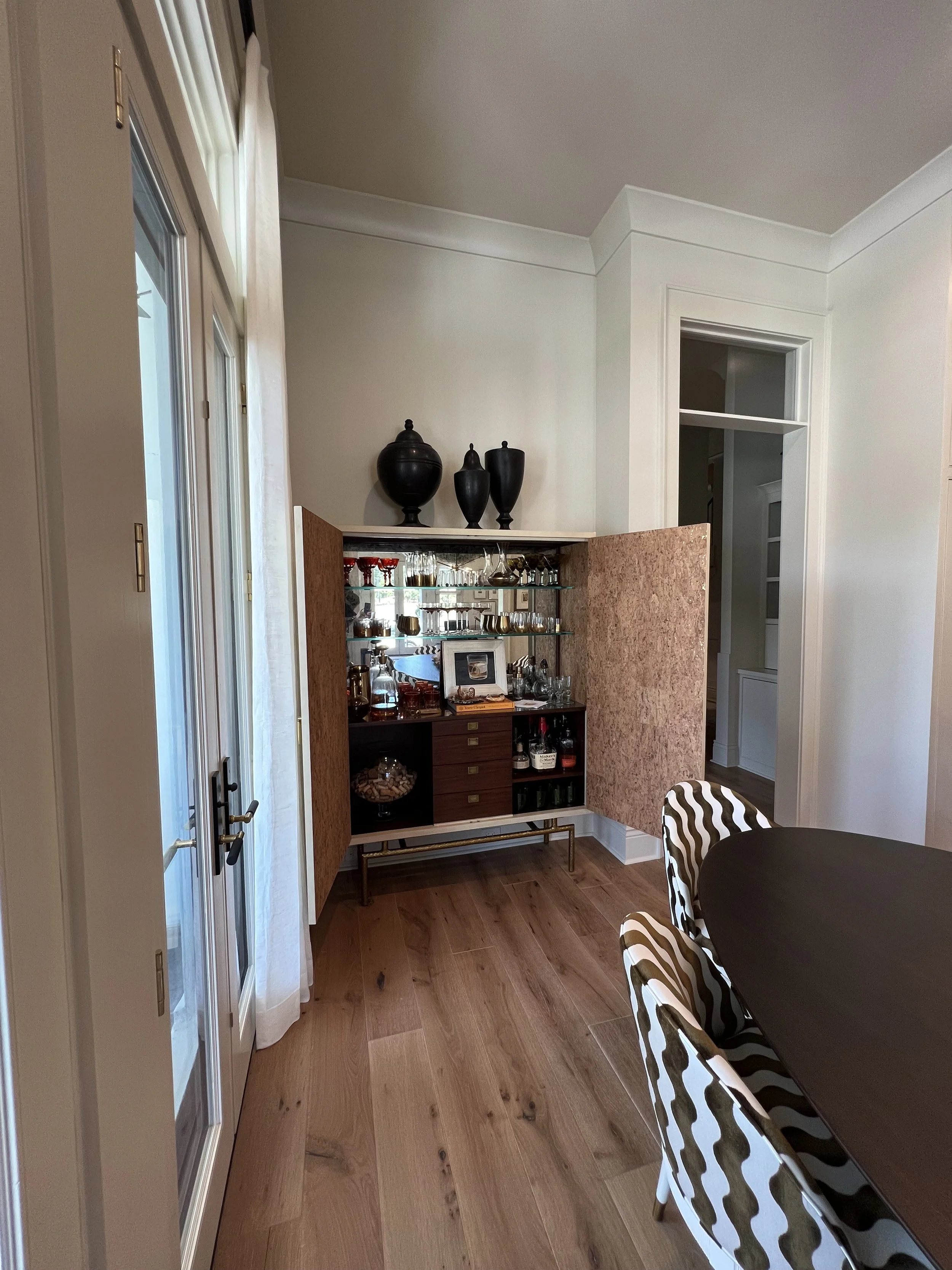 A corner of a dining room with a wooden cabinet filled with glassware and bottles. On top of the cabinet are three black vases. The room has wooden flooring, a black dining table, and chairs with striped upholstery. French doors with white curtains l