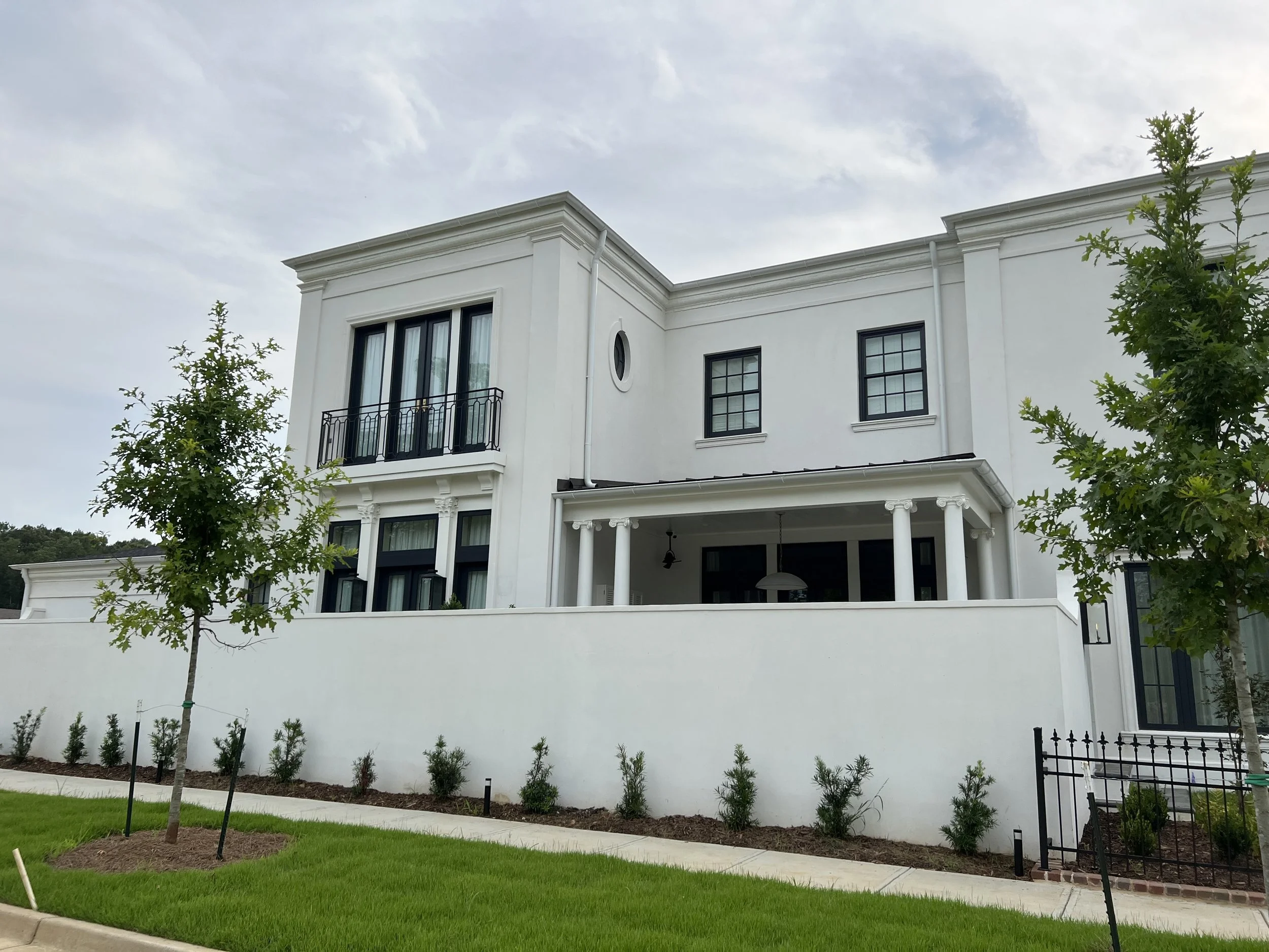 White modern multi-story house with large windows, a balcony, and a front yard with small trees and a manicured lawn.