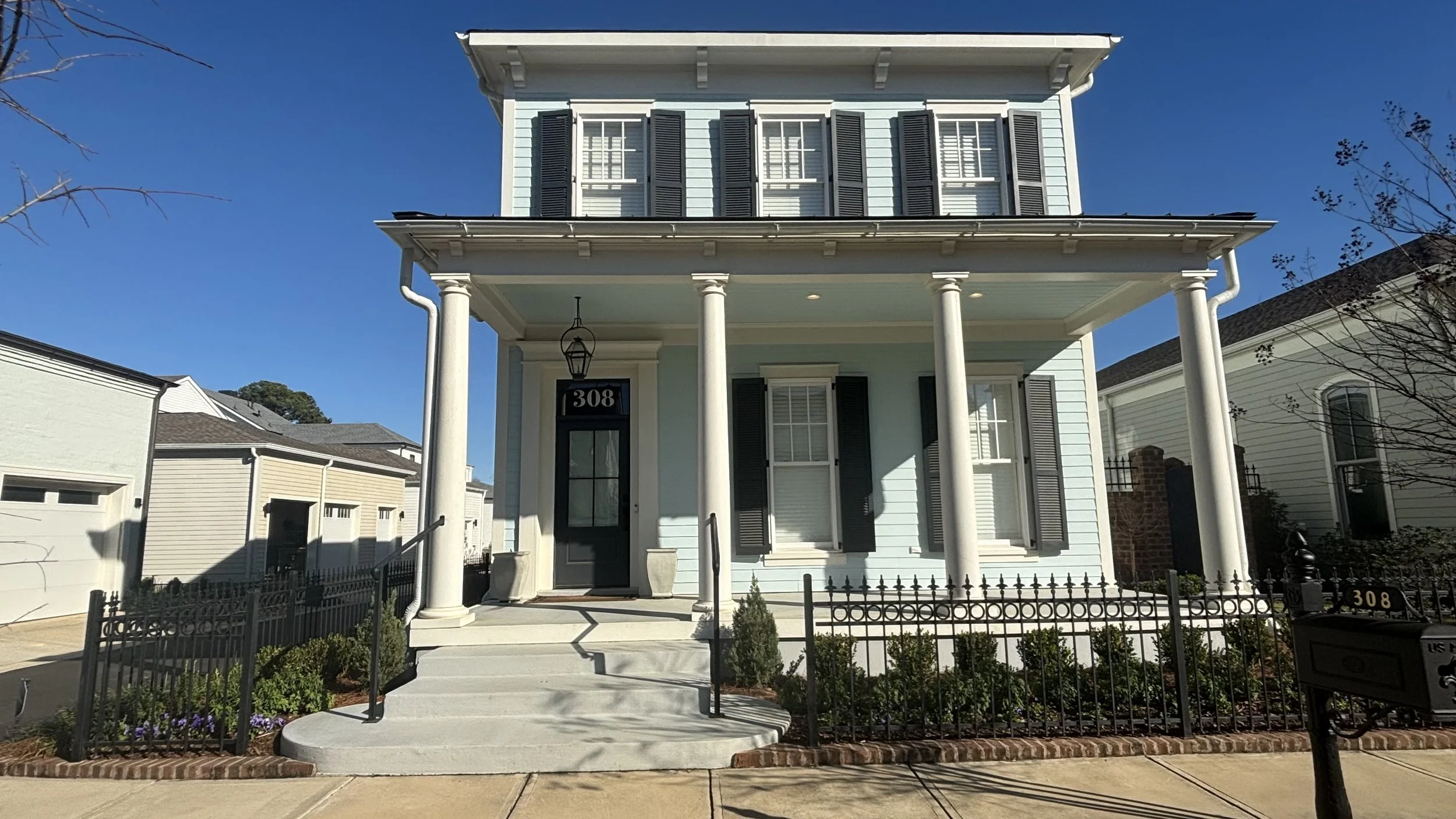 A two-story light blue house with white columns and black shutters, featuring a front porch and steps leading up to the door, with a small garden and black fence in front.