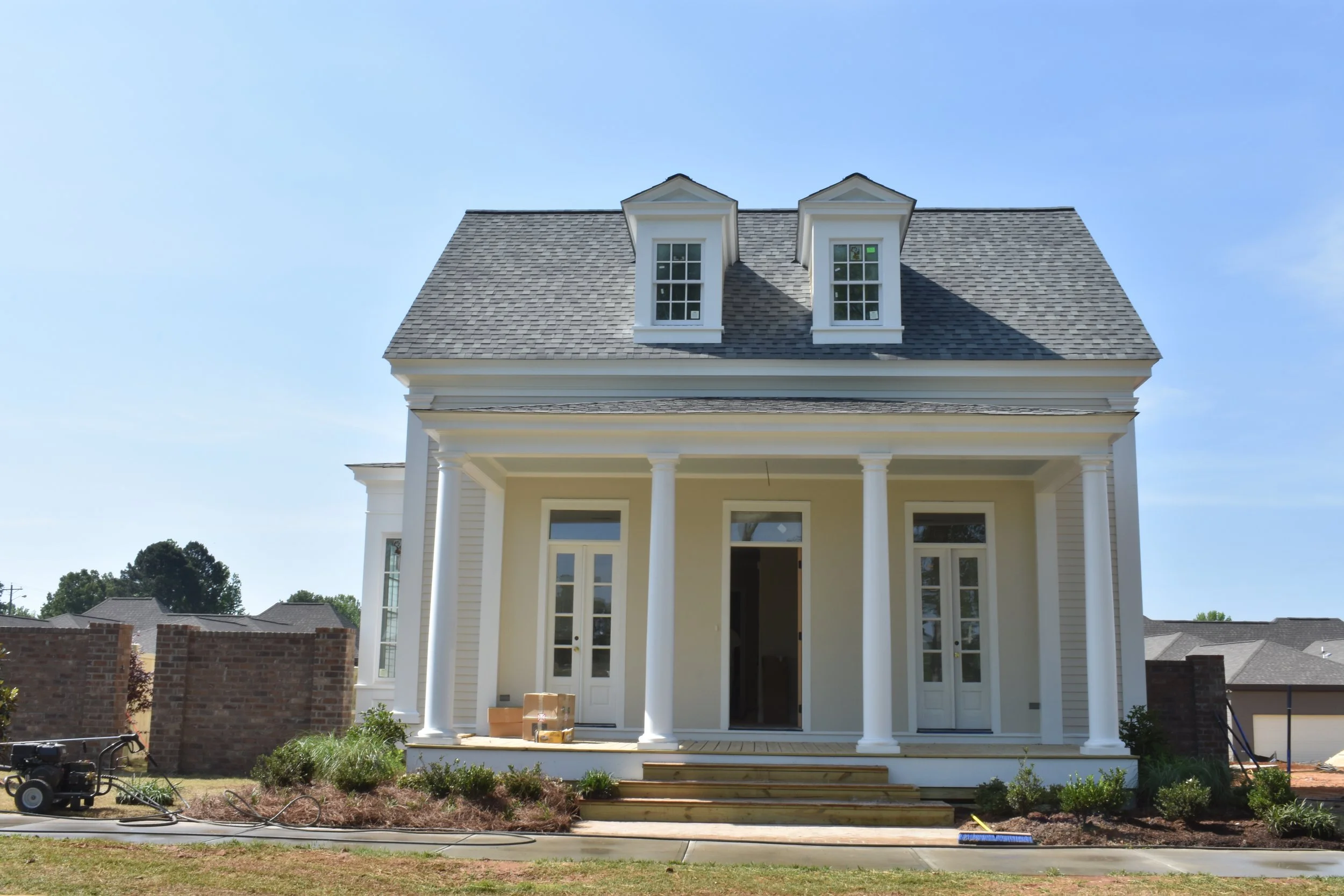 Front view of a new, white, two-story house under construction with columns on the porch and two dormer windows on the roof, with some construction materials and tools visible in the yard.