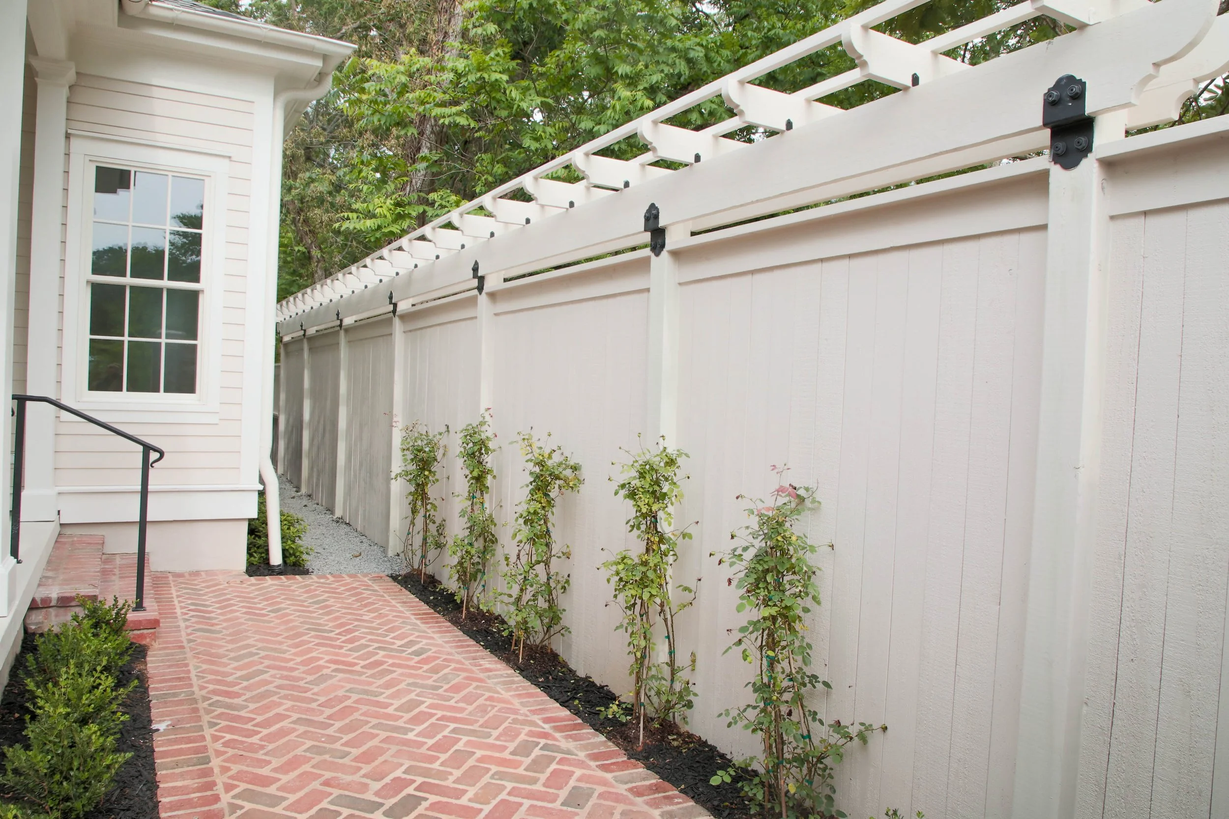A backyard with a white wooden privacy fence, a brick patio, and young plants growing along the fence line next to a house with a window and a set of steps.