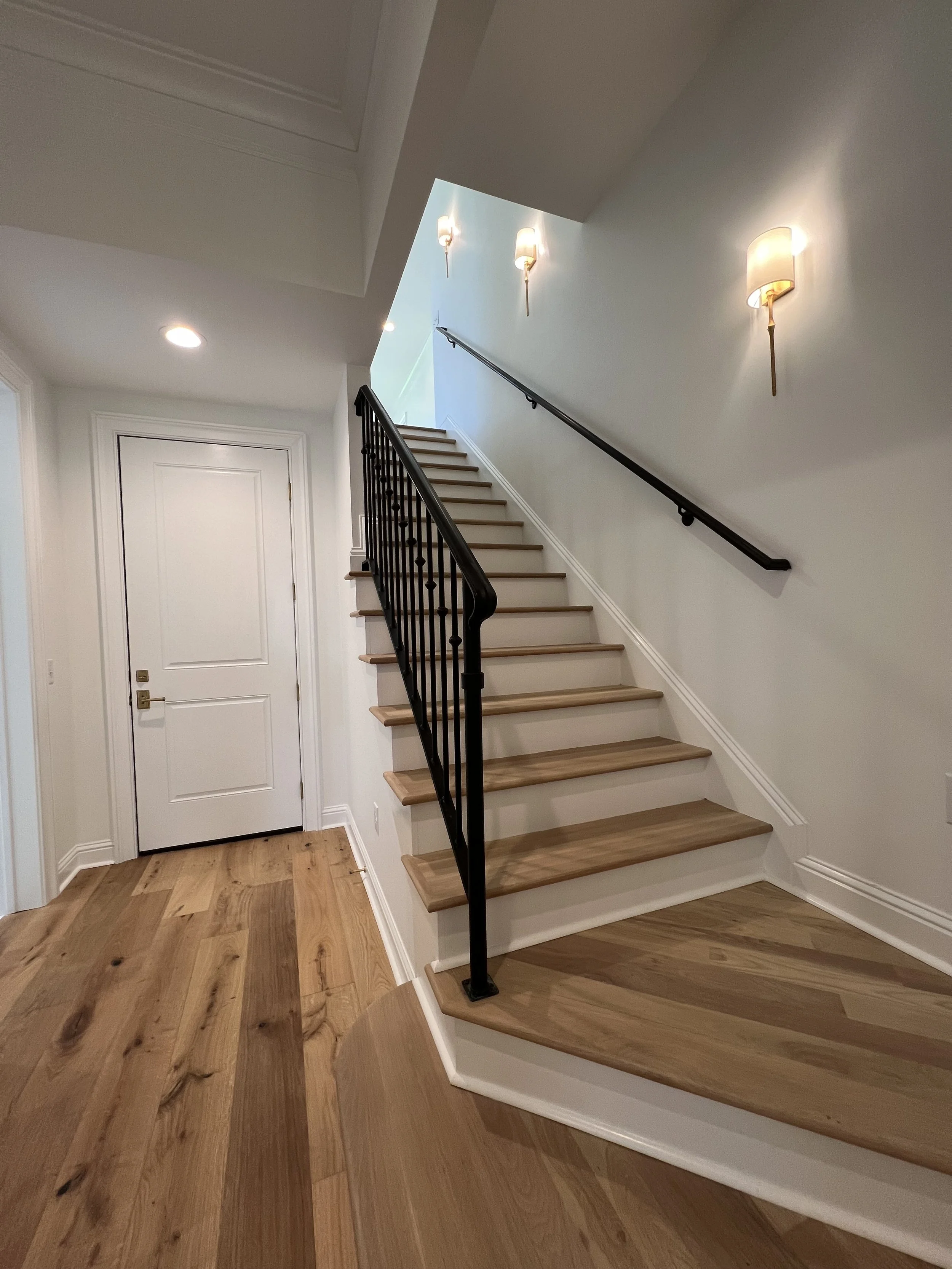 Interior view of a staircase with wooden steps, black metal railing, and wall-mounted lights, leading upward inside a residential home.