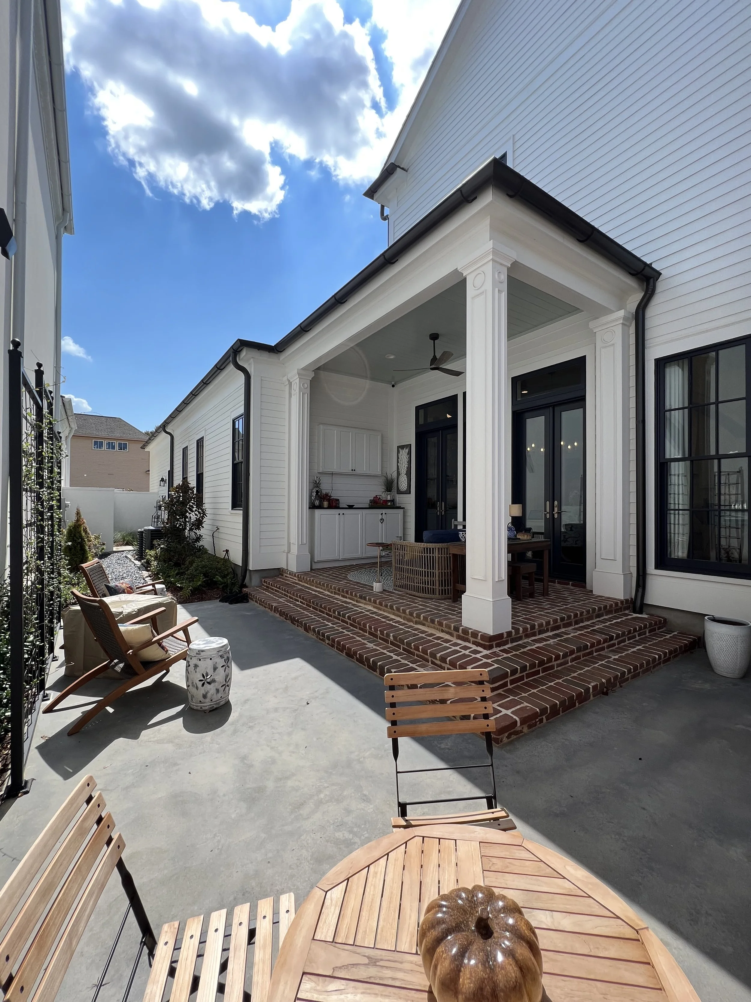Backyard patio with brick steps, outdoor seating including chairs and tables, and a white house with black window frames and a covered porch.