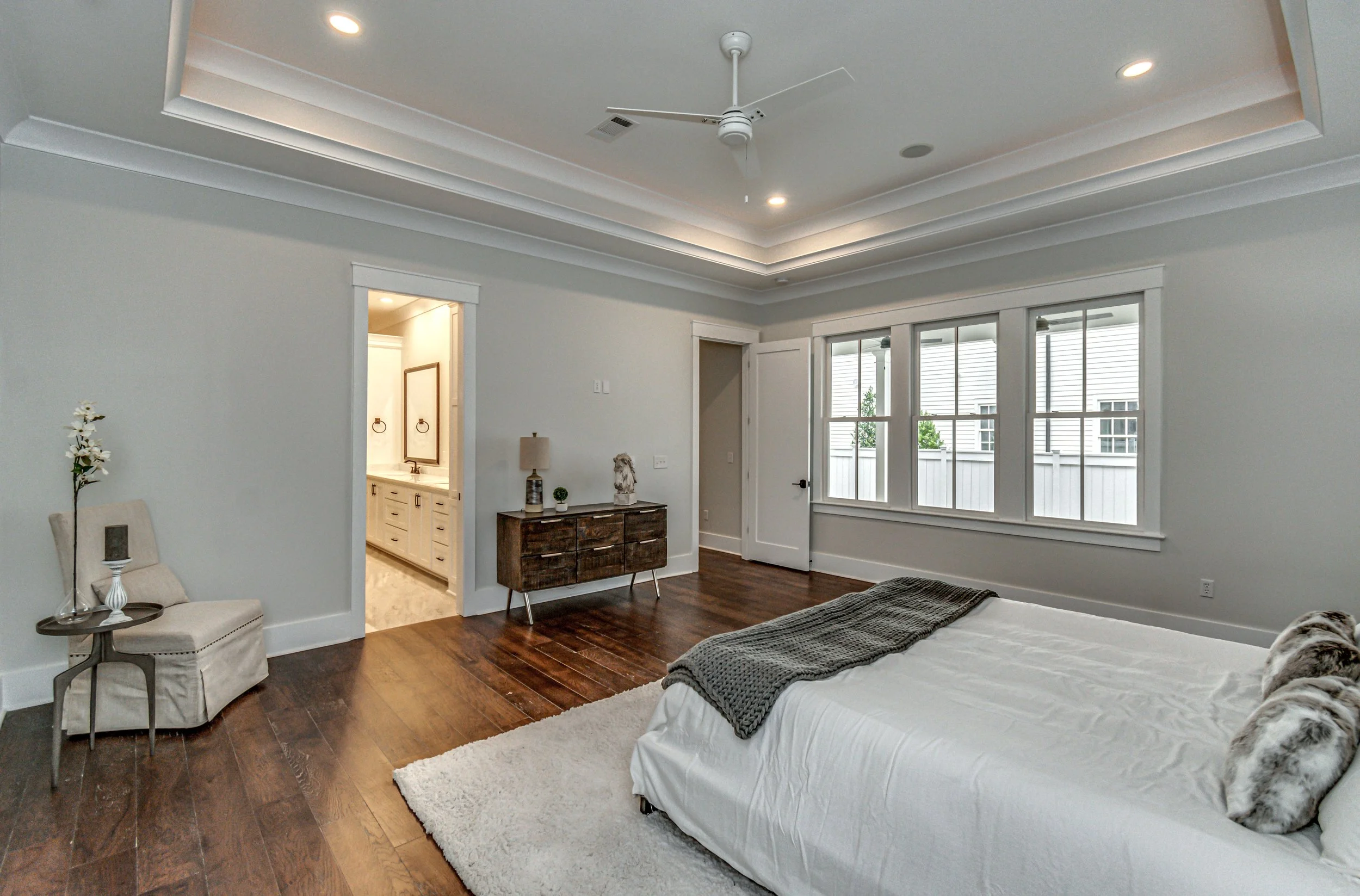 A spacious bedroom with a large bed, wooden floors, a white rug, and three fluffy pillows. There is a chair with a flower and a small framed picture, a dark wooden dresser with decor items, and a window with white trim overlooking an exterior area. T