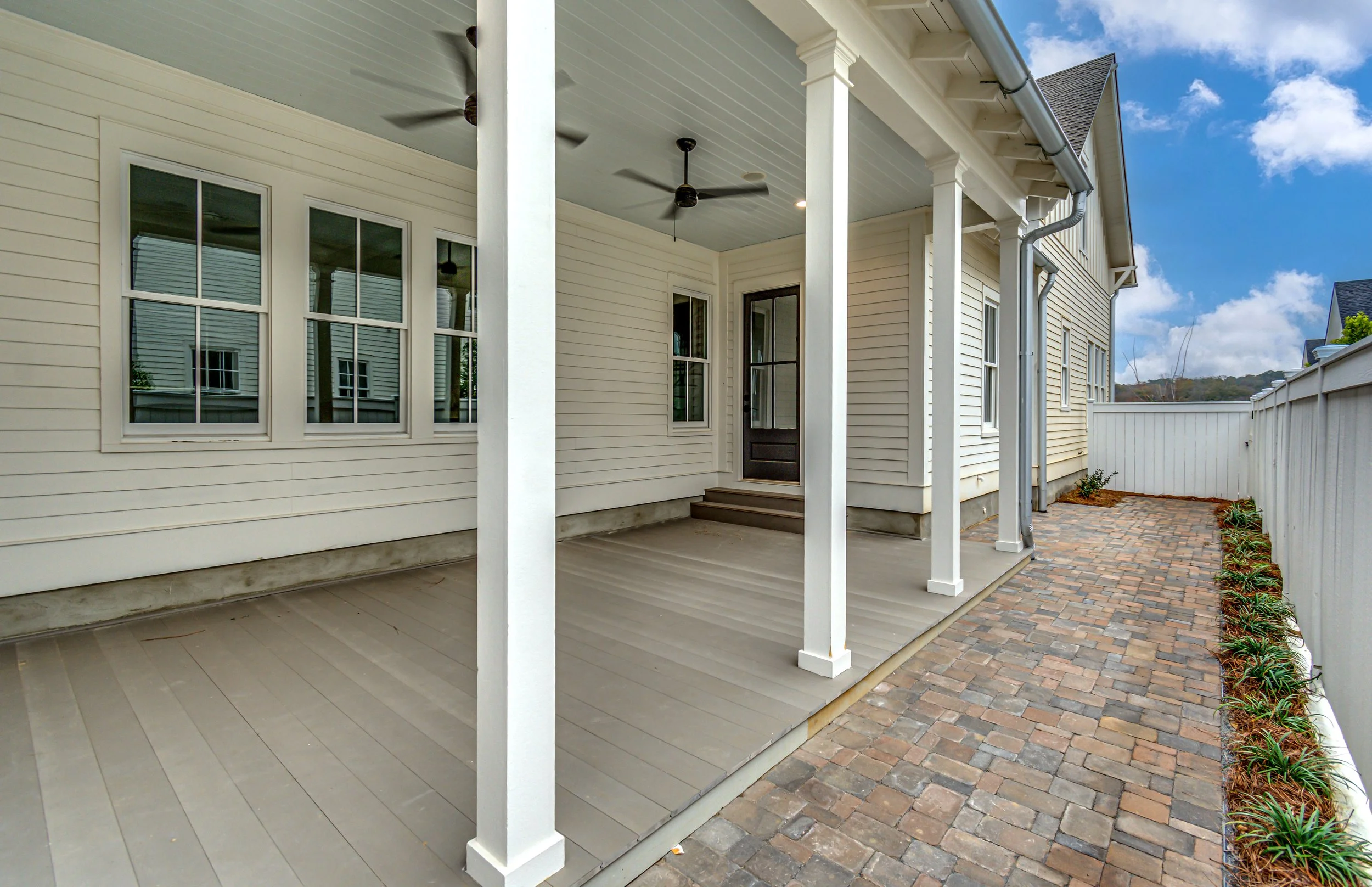 Back porch area with beige flooring, white support columns, ceiling fans, a door, and a brick pathway along the white fence with plants.