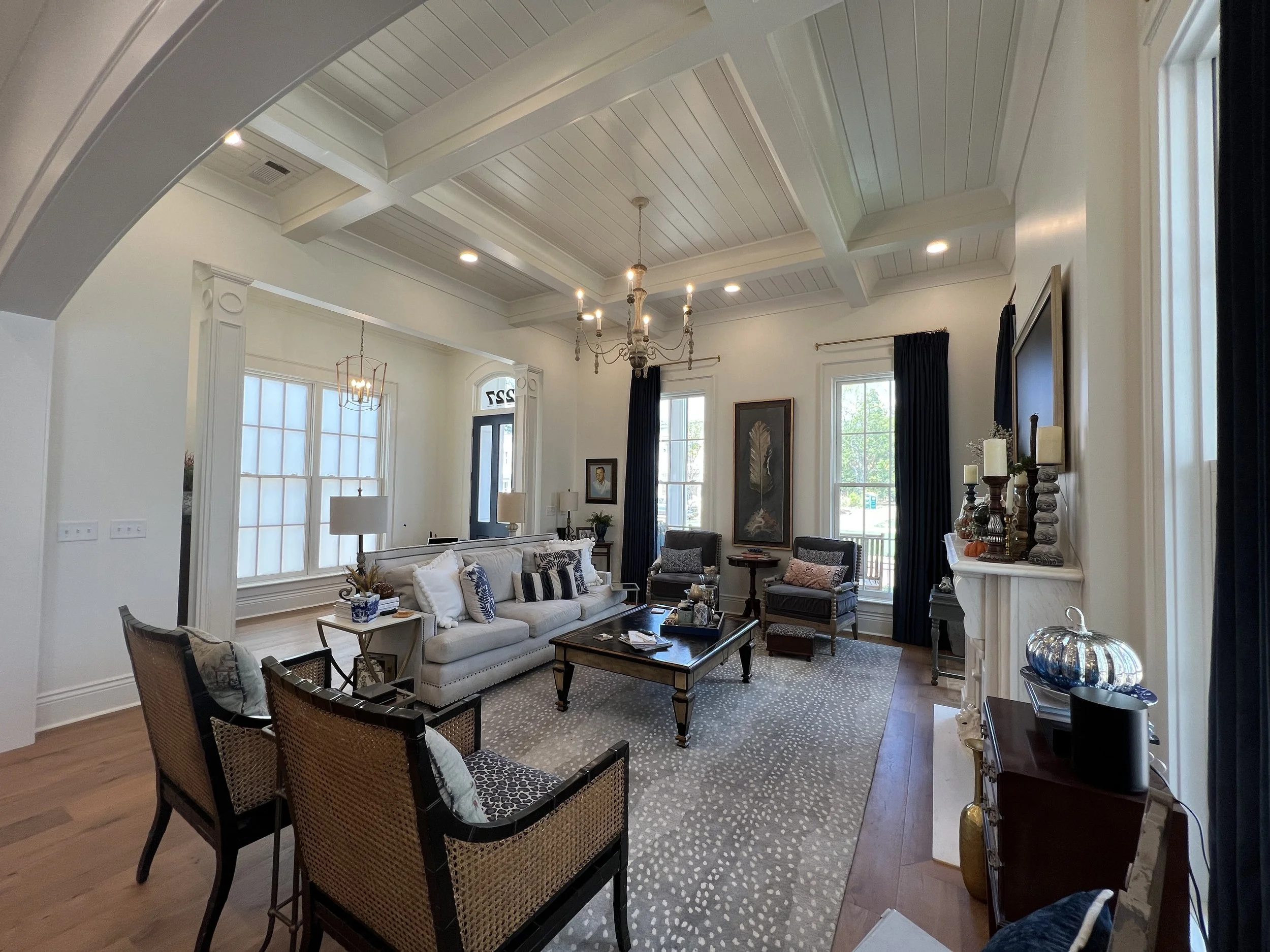 Living room with white walls, hardwood floors, large windows with dark curtains, a white sofa, armchairs, a coffee table, and various decorative items.
