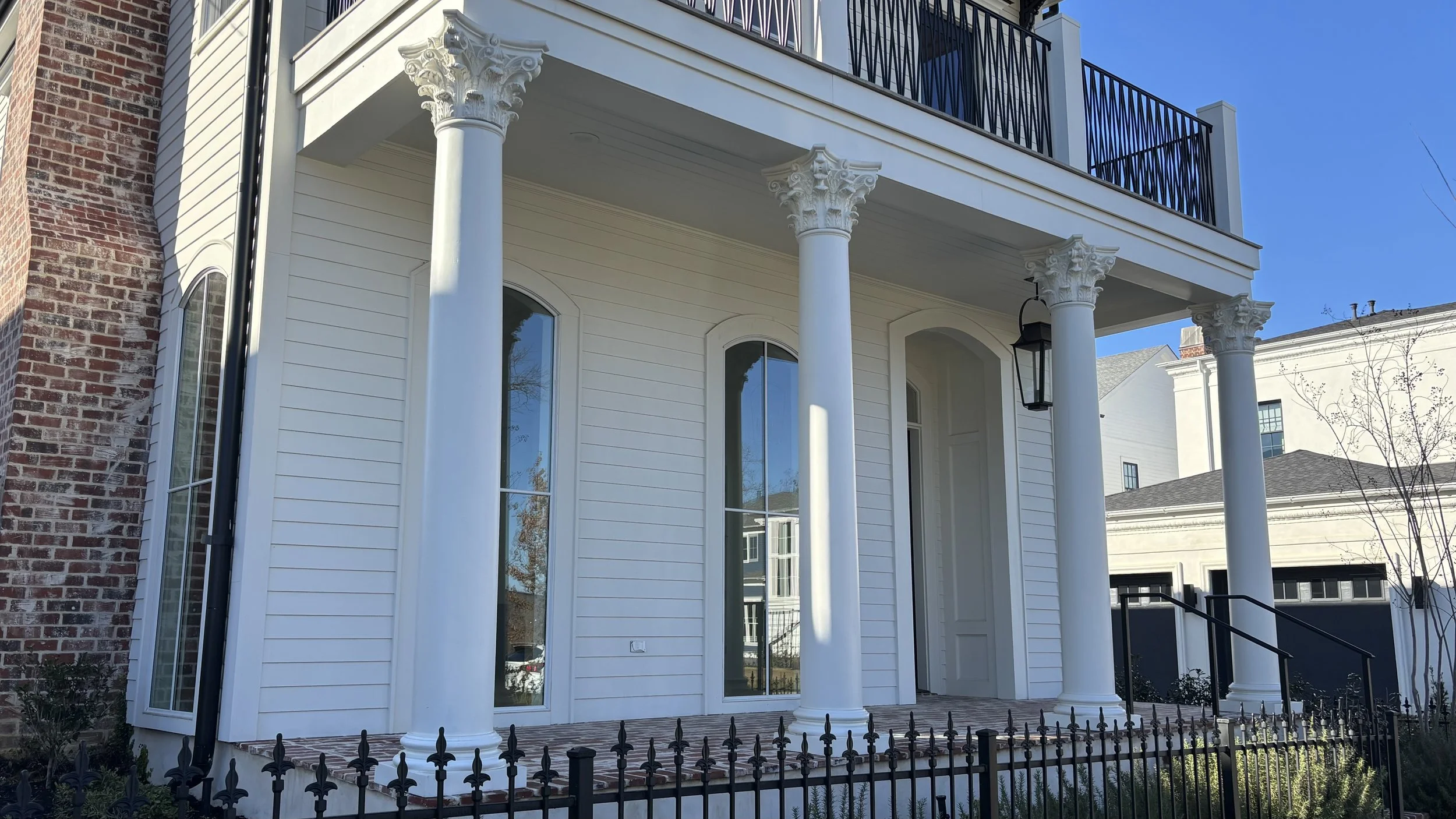 Front porch of a house with four white columns with decorative capitals, black metal railing on upper balcony, black outdoor lantern, large arched windows, brick and white siding exterior, and small landscaped yard with black fence.
