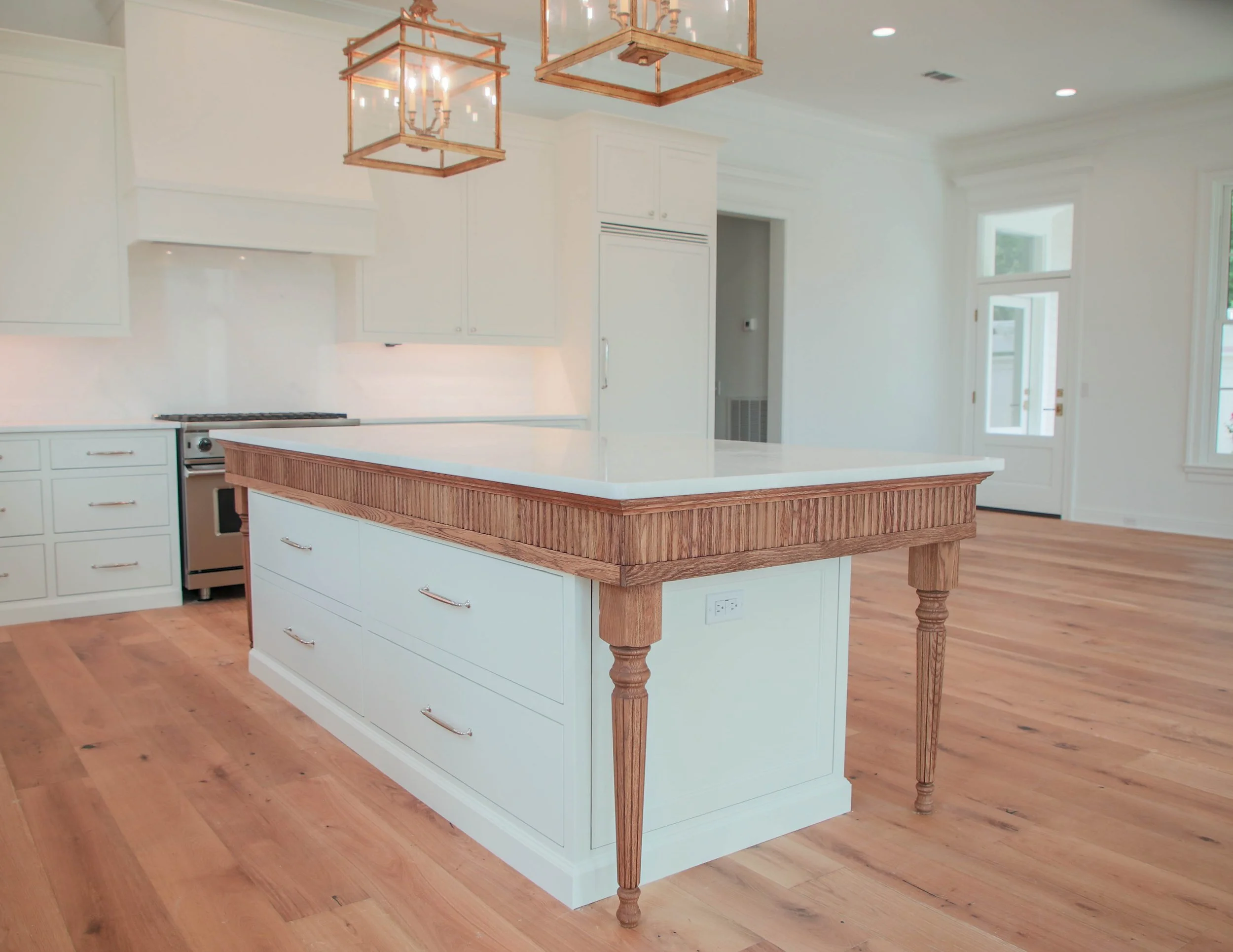 Empty kitchen with a large island featuring a white countertop and wood accents, surrounded by white cabinets and hardwood floors, with large windows and modern lighting fixtures.