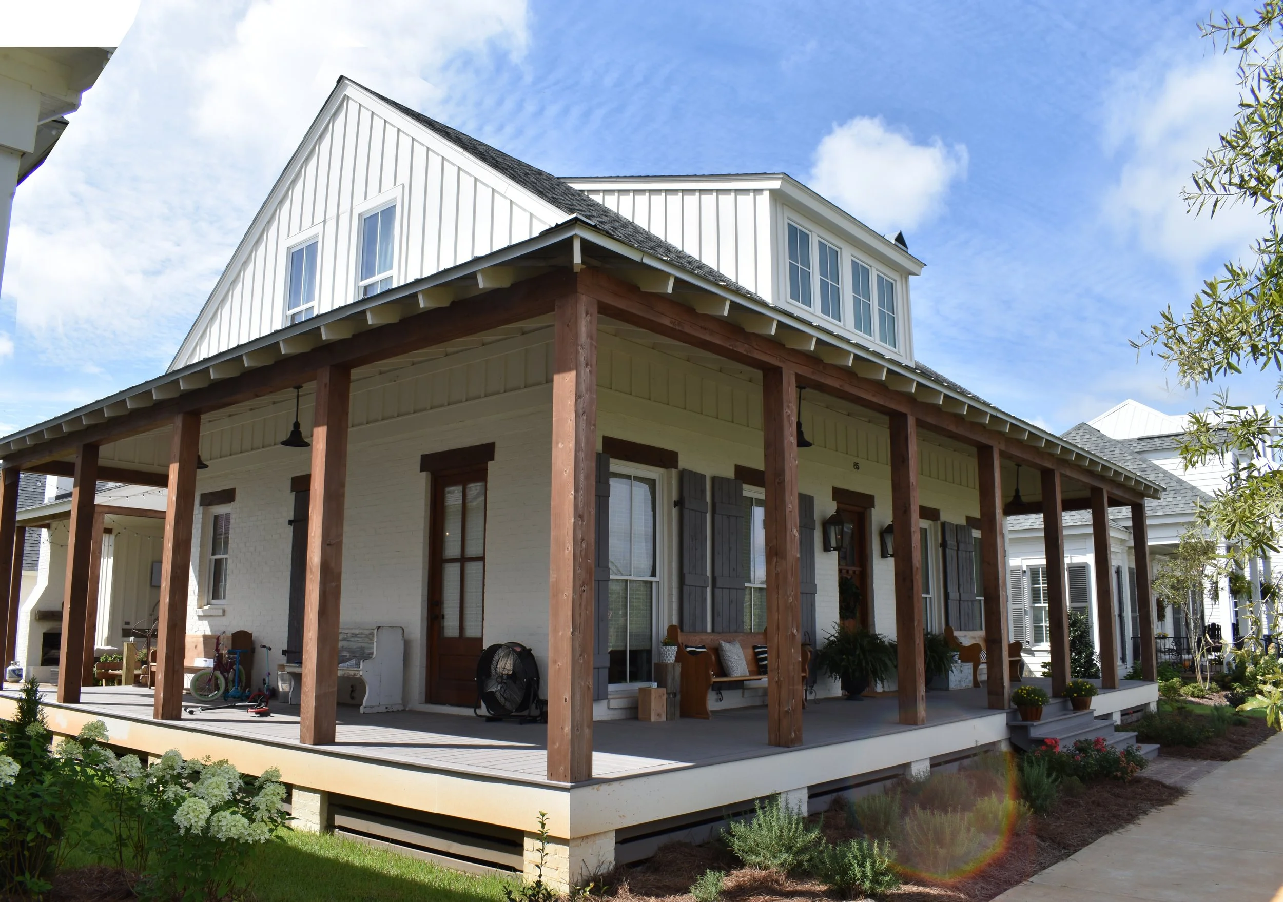 A two-story house with a large front porch, white siding, brown shutters, and a gray shingle roof under a partly cloudy sky. The porch has wooden support beams and various outdoor furniture.