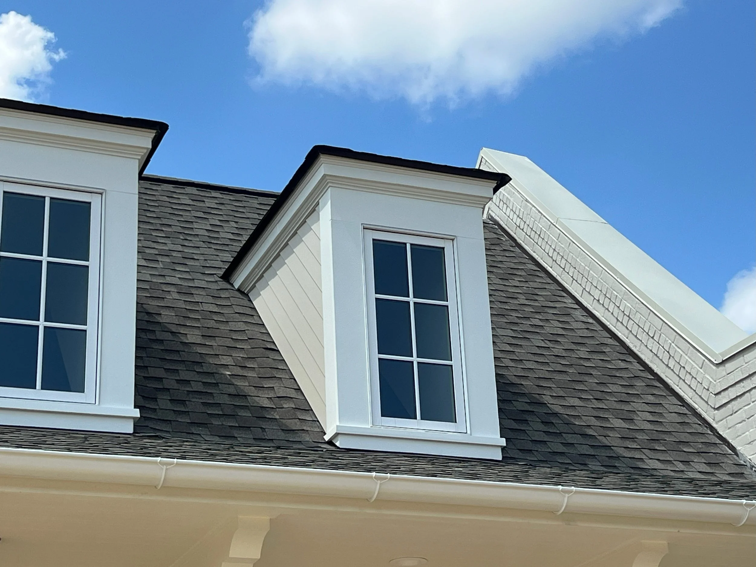 Close-up of a house's exterior featuring a steep roof with gray shingles, white window frames, and dormer windows set against a blue sky with scattered clouds.
