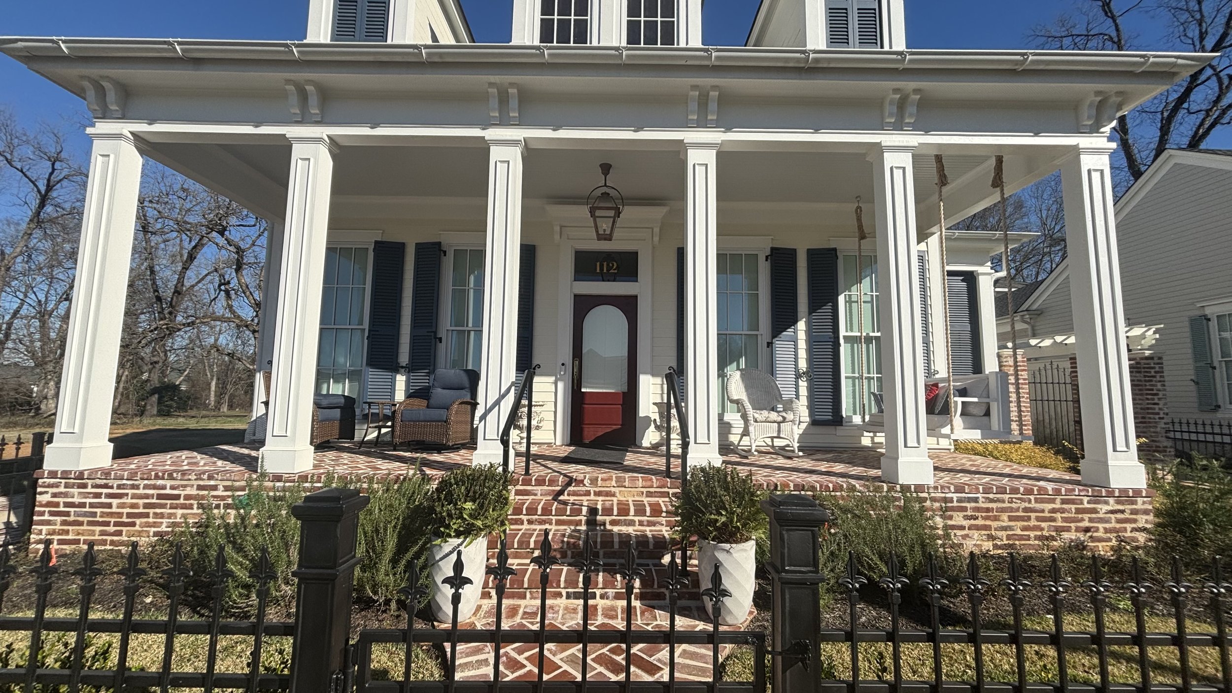 Front porch of a house with white columns, black shutters, wicker and metal chairs, and a brick walkway and stairs.