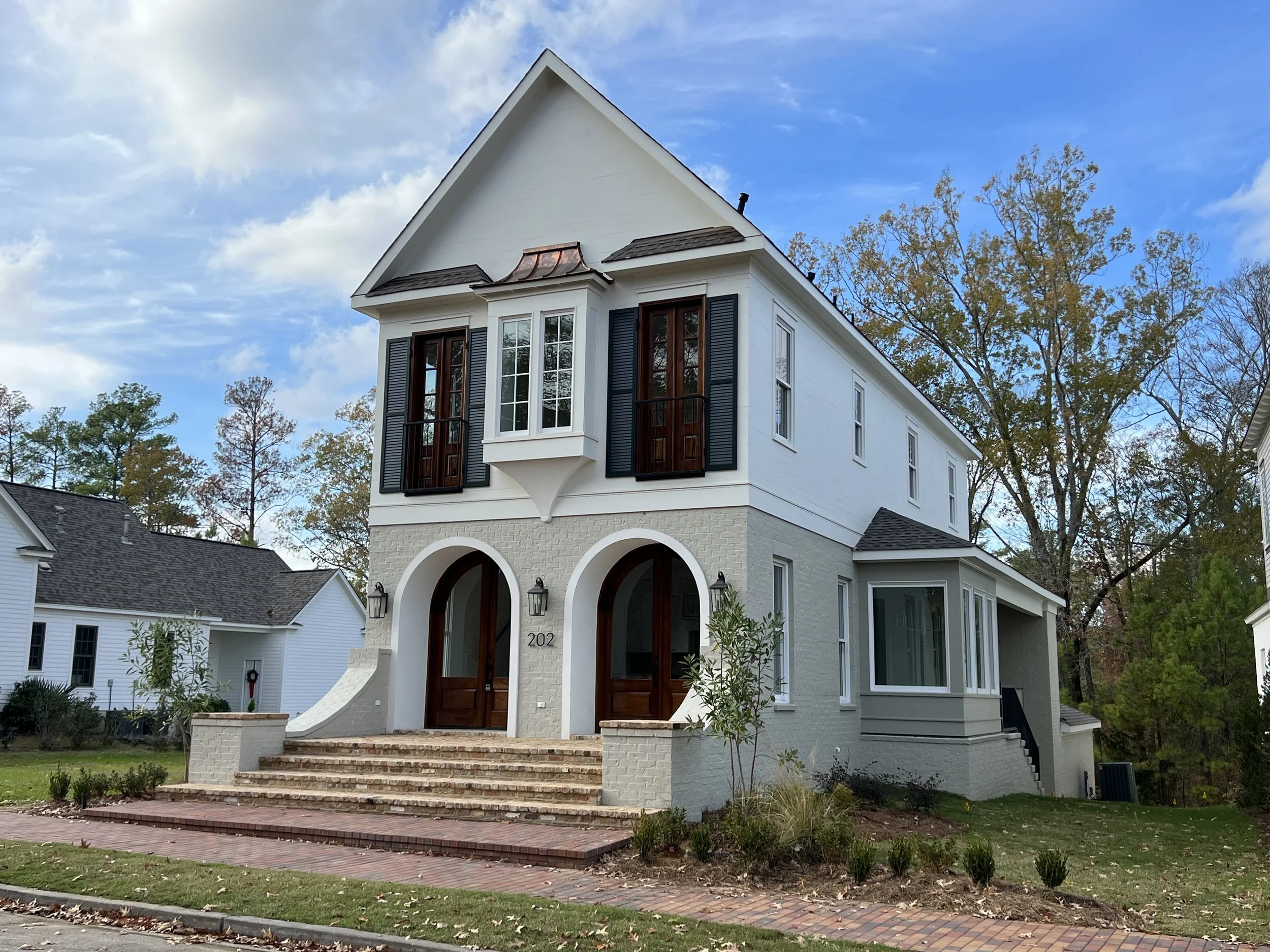 A modern two-story house with white brick and siding exterior, black shutters, arched front doors, and steps leading to a brick walkway, surrounded by trees and a well-maintained lawn.