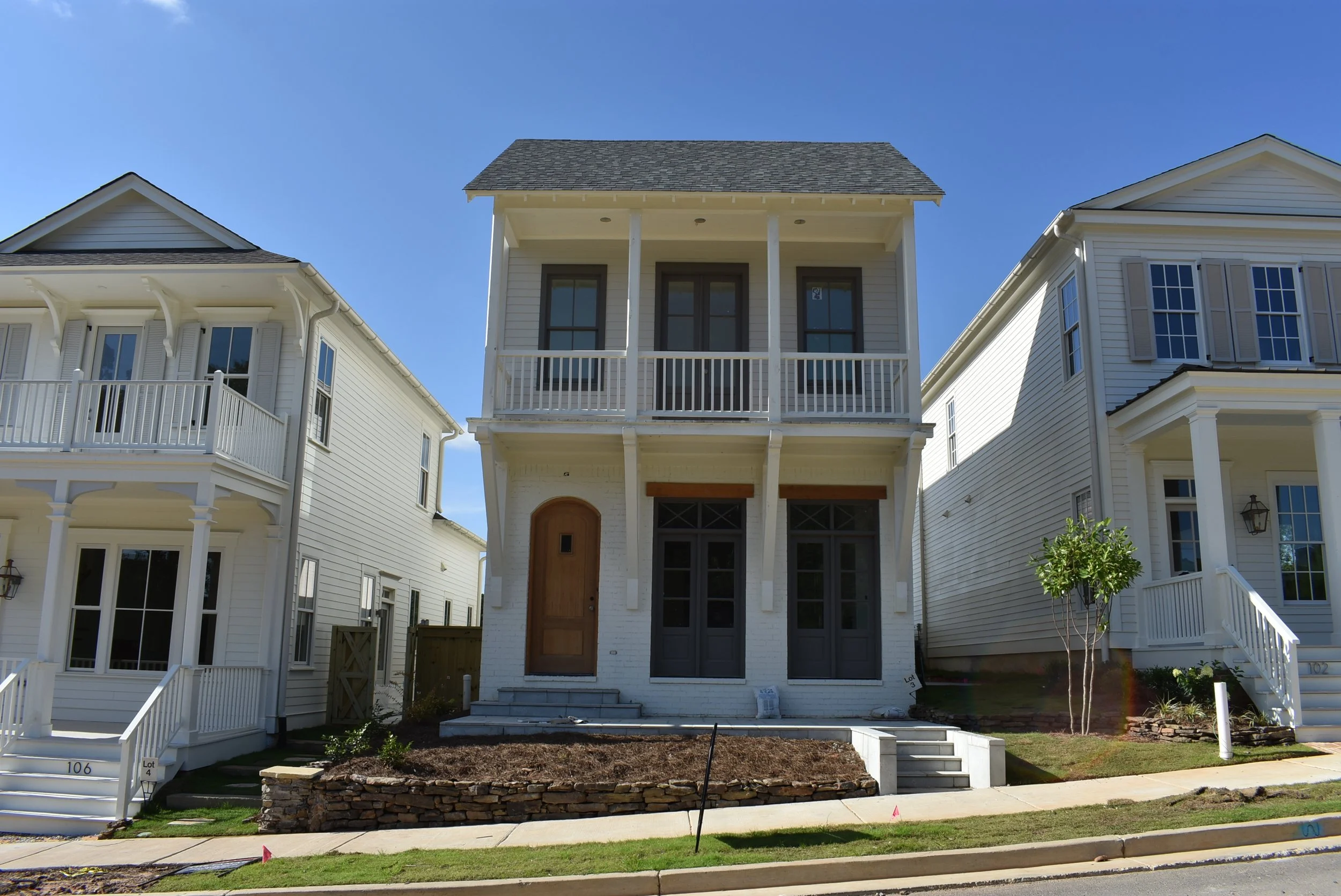 Newly constructed multi-story house with white siding, a small front porch, and steps leading up to the entrance. The house has three large windows on the front and a balcony on the second floor. The neighboring houses are similar in style and color,