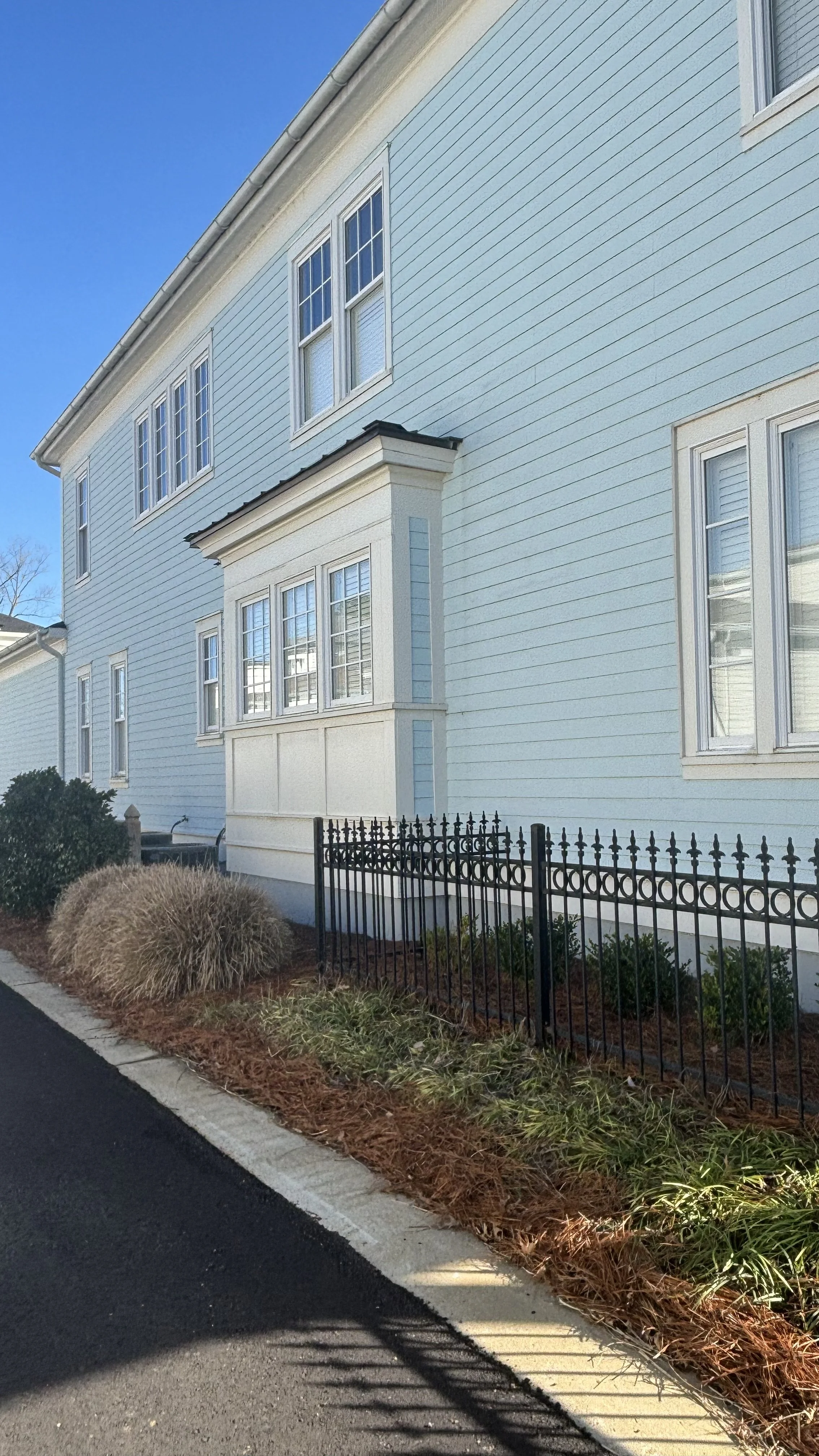 A light blue, multi-story house with white trim, multiple windows, and a small garden with shrubs, surrounded by a black metal fence, under a clear blue sky.