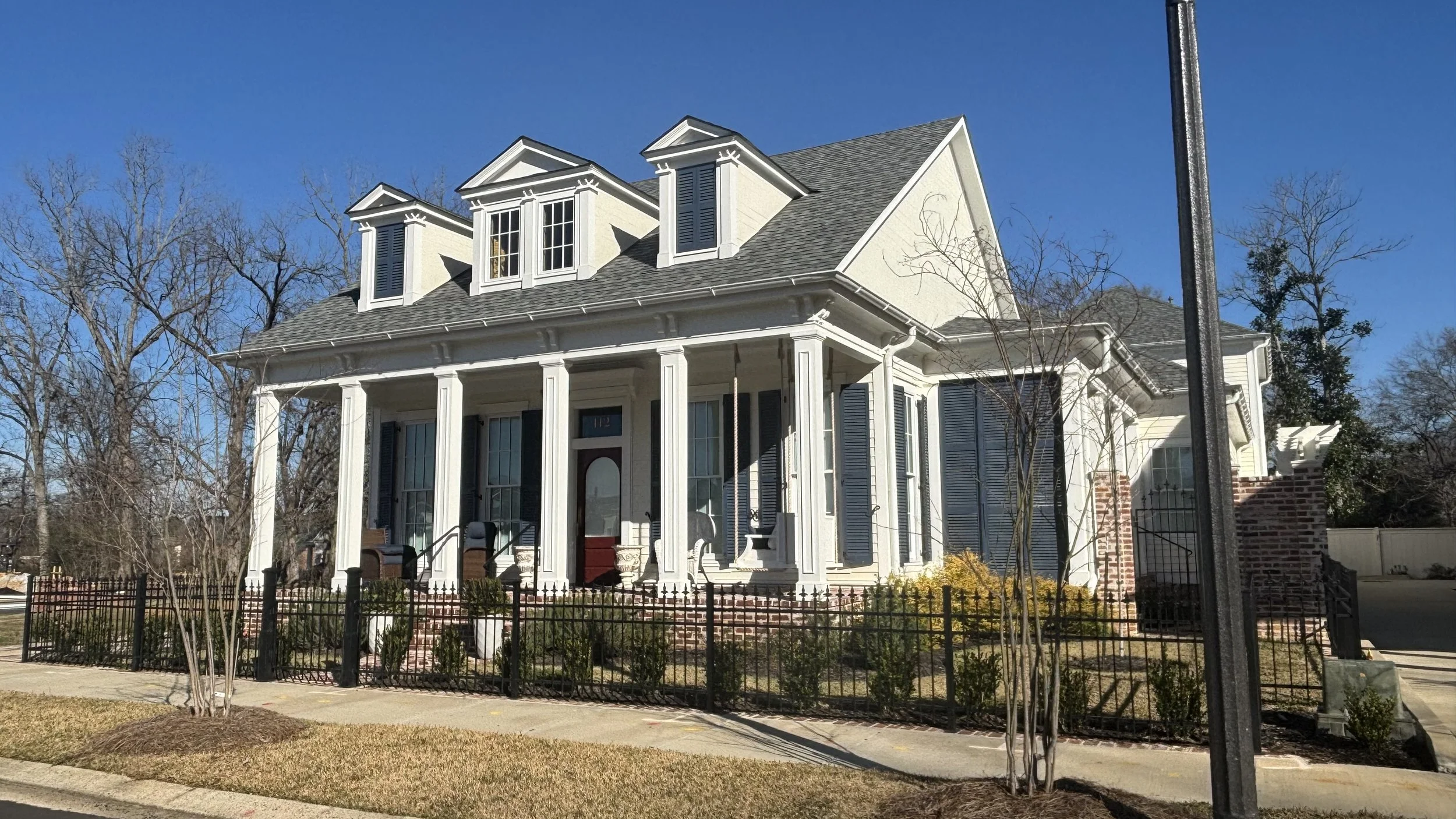 Large white house with a porch, multiple windows, and dormer windows on the roof. Front yard has small bushes and trees, black iron fence, and a sidewalk. Clear blue sky.
